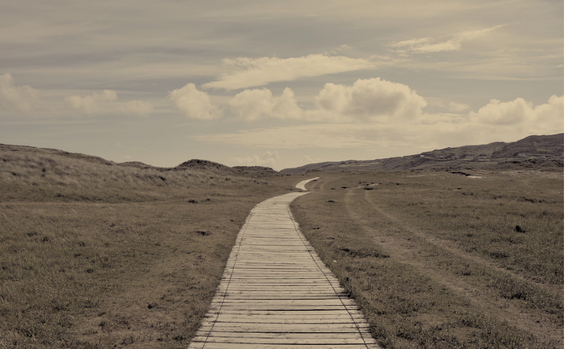 A path winding through an open grassy field with hills in the distance and some clouds overhead. Suggests a journey.