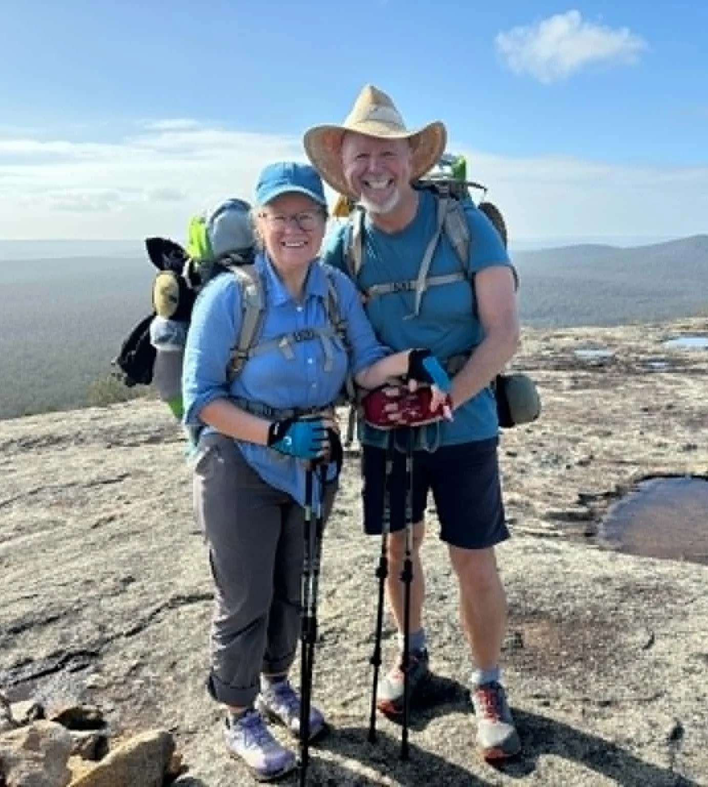 Two hikers, a woman and a man, standing together outdoors on a rocky terrain with a clear sky in the background. They are smiling and wearing hats, backpacks, and hiking gear.