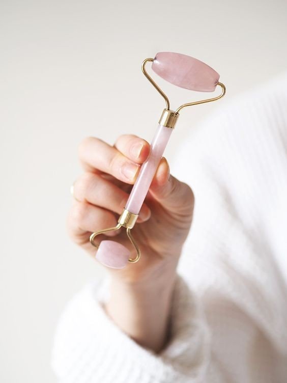 A hand holding a pink jade facial roller with gold accents.