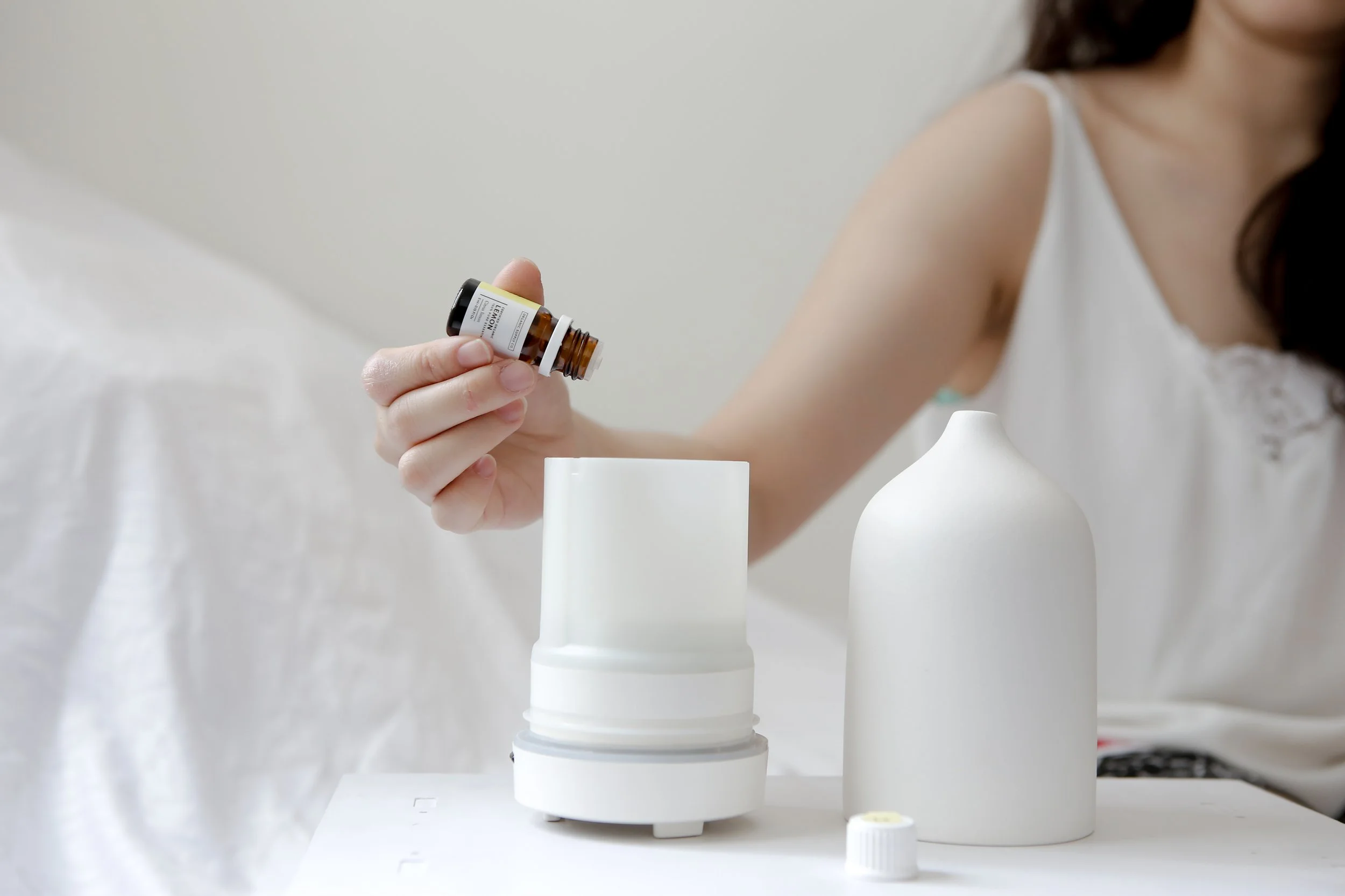 A person holding a small brown bottle labeled lemon essential oil, with two white bottles on a white surface, and a white cylindrical object beneath them.