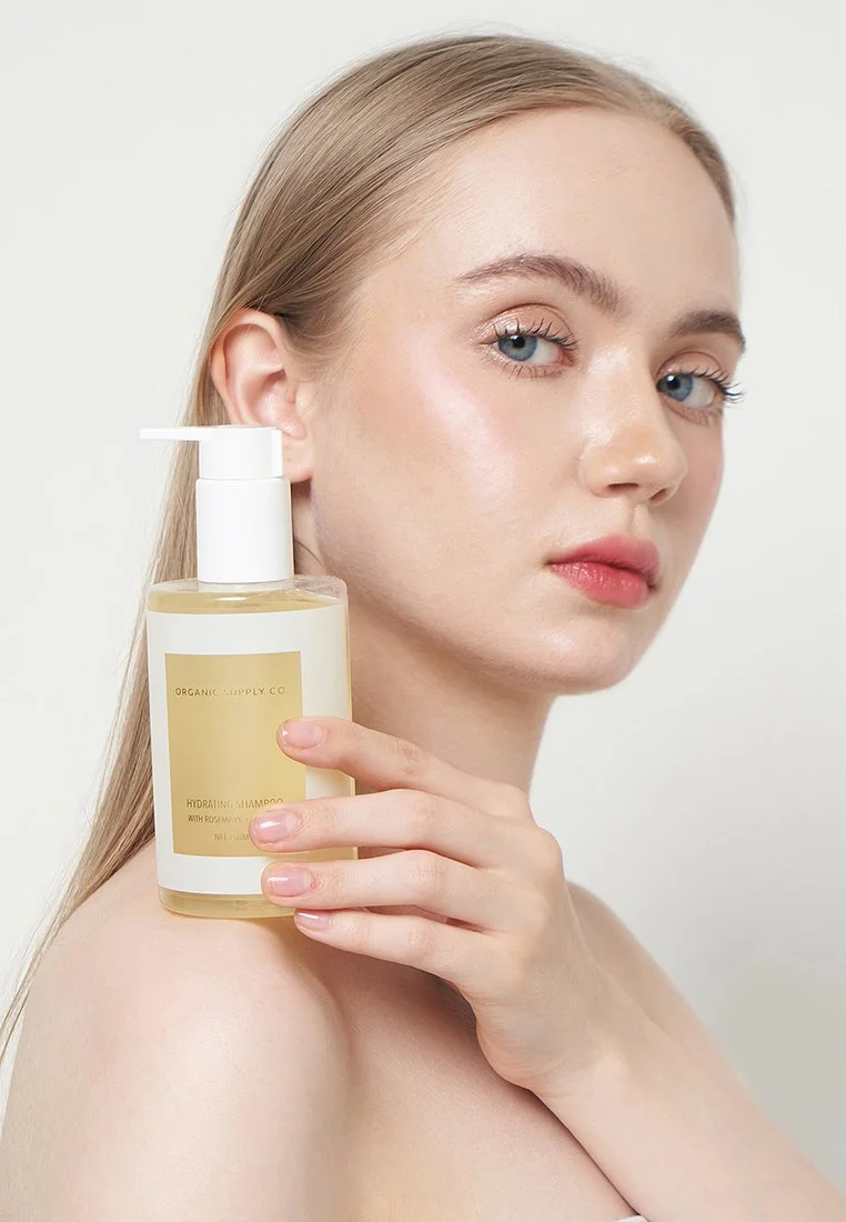 A young woman with fair skin and blue eyes holding a bottle of organic hydrating shampoo with rosehip and coconut oil.