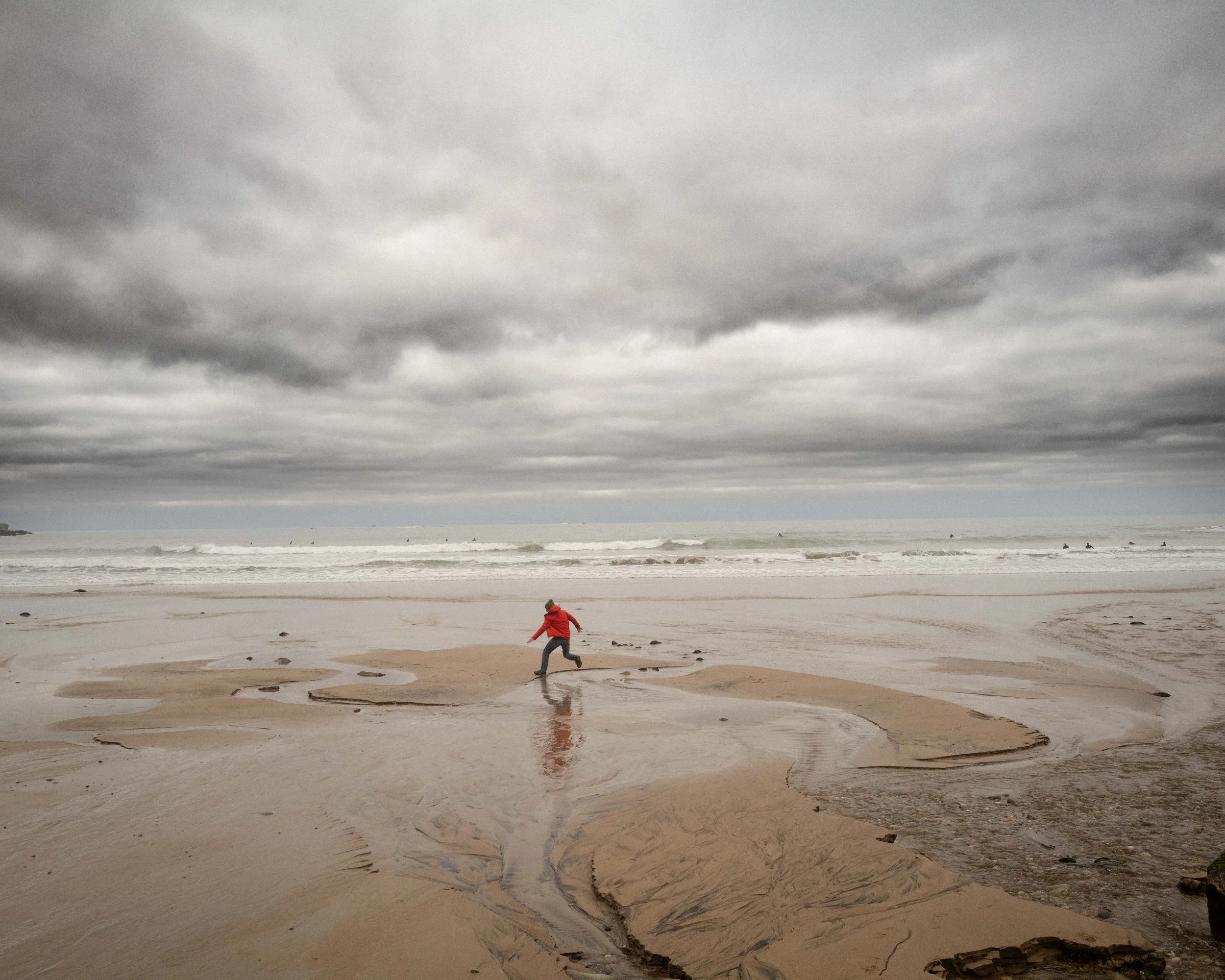A person in a red jacket running along a beach with dark storm clouds overhead and a few surfers in the water.