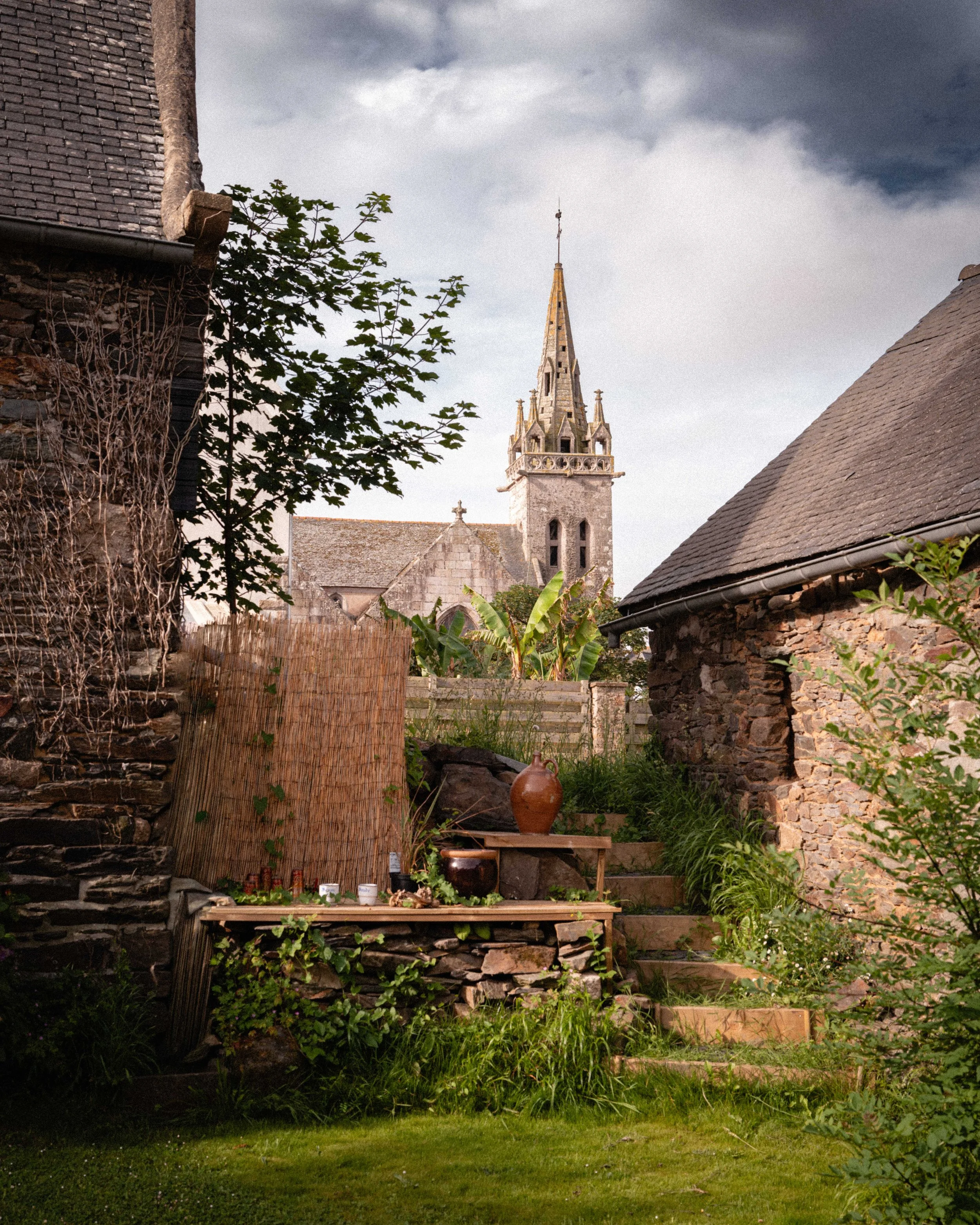 A backyard with stone and brick buildings, a grassy lawn, plants, and a stone church with a tall, pointed steeple in the background under cloudy sky.