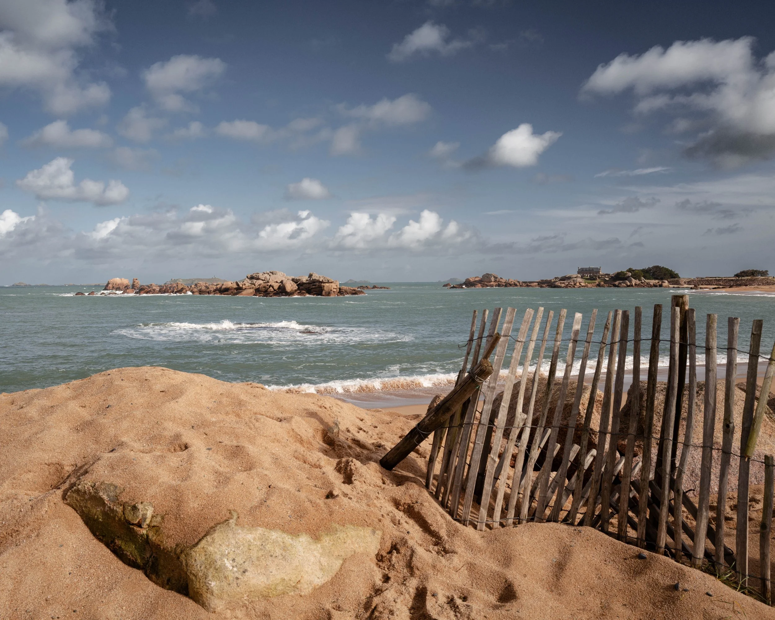 A sandy beach with rocks and a wooden fence in the foreground, small islands with houses and rocky outcroppings in the background, and a partly cloudy sky.