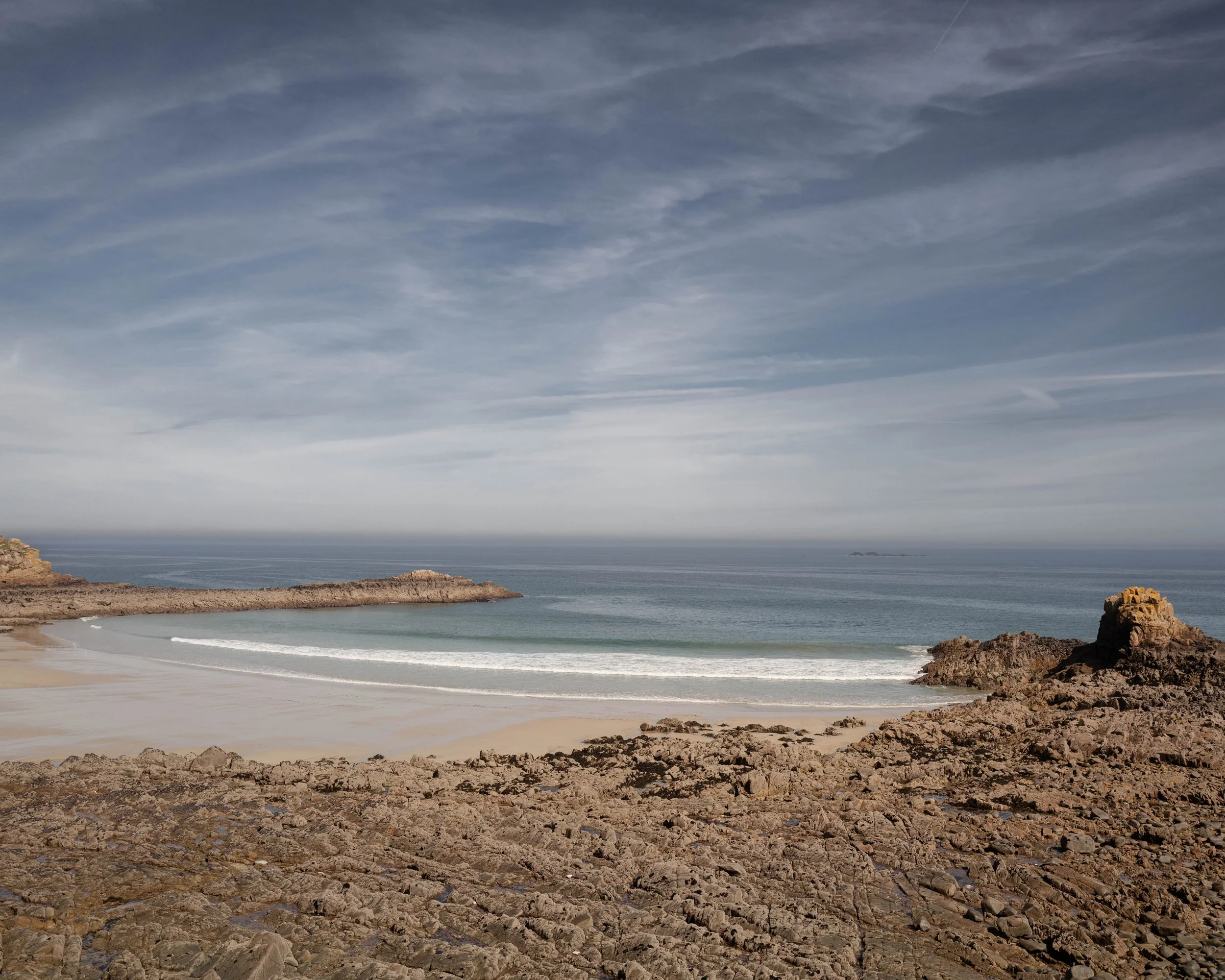 A rocky coastline with a sandy beach and calm ocean waves under a cloudy sky.