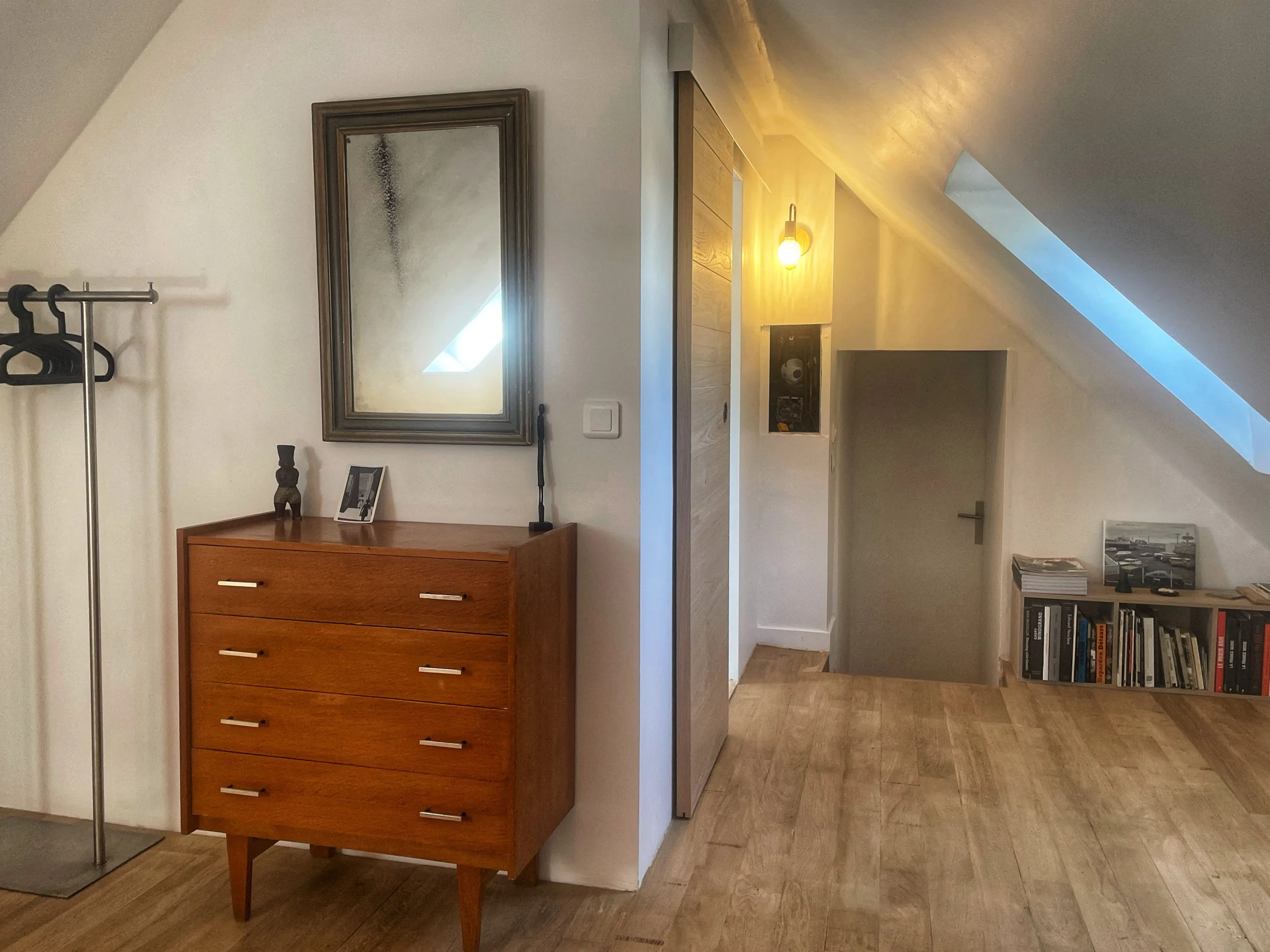A room with sloped ceilings, a wooden dresser, a mirror, a coat rack, a small table with books, a bookshelf, and a skylight window.
