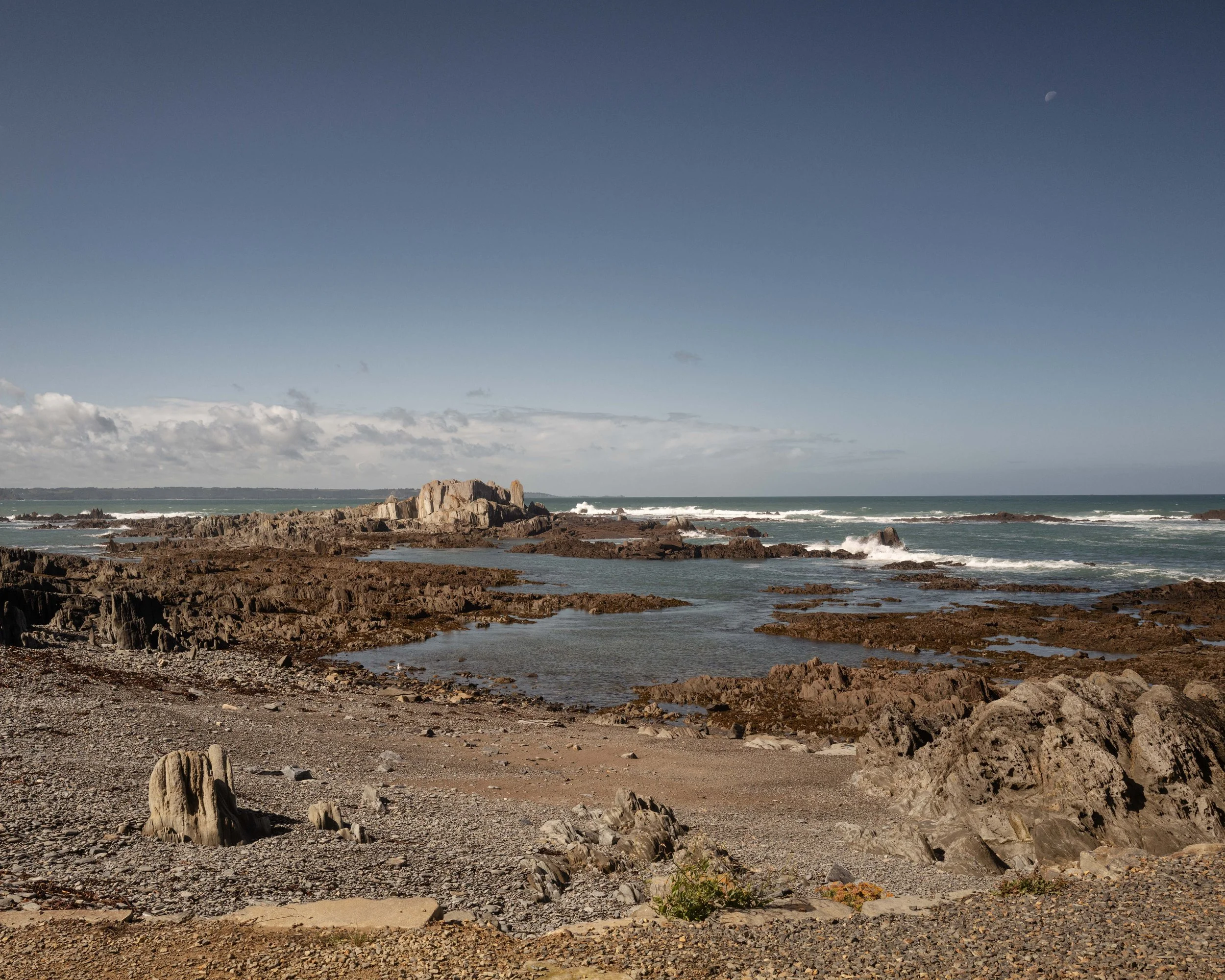 Coastal scene with rocky shoreline, ocean waves, a large rock formation, cloudy sky, and a visible moon.