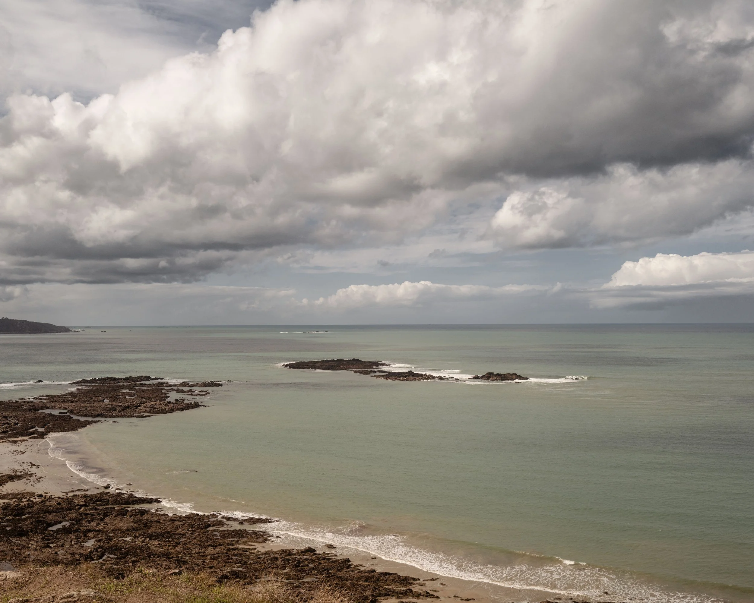 Overcast sky with dark clouds over a calm ocean with rocky shoreline in the foreground.
