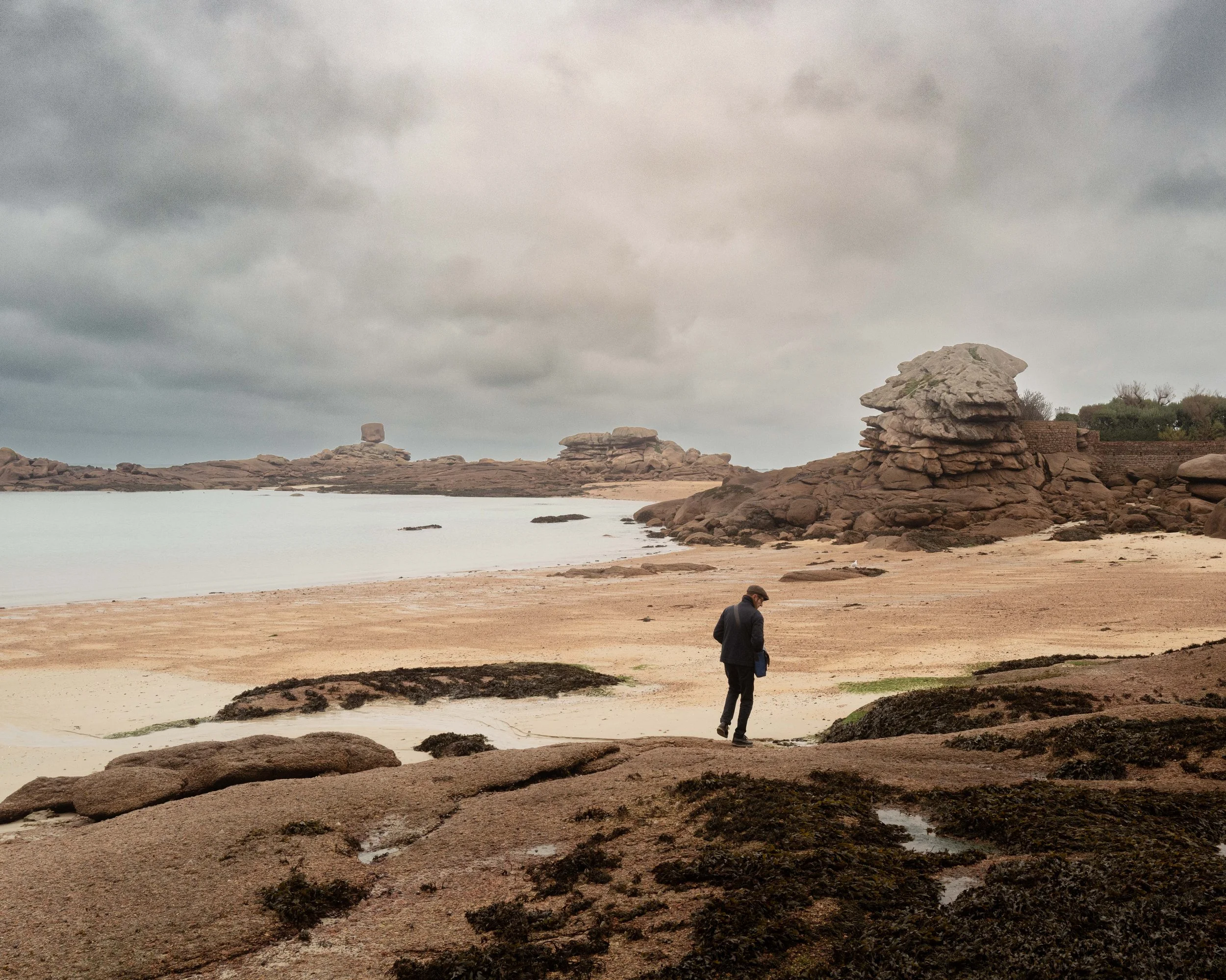 A person walking along a rocky beach under a cloudy sky with large rocks and water in the background.