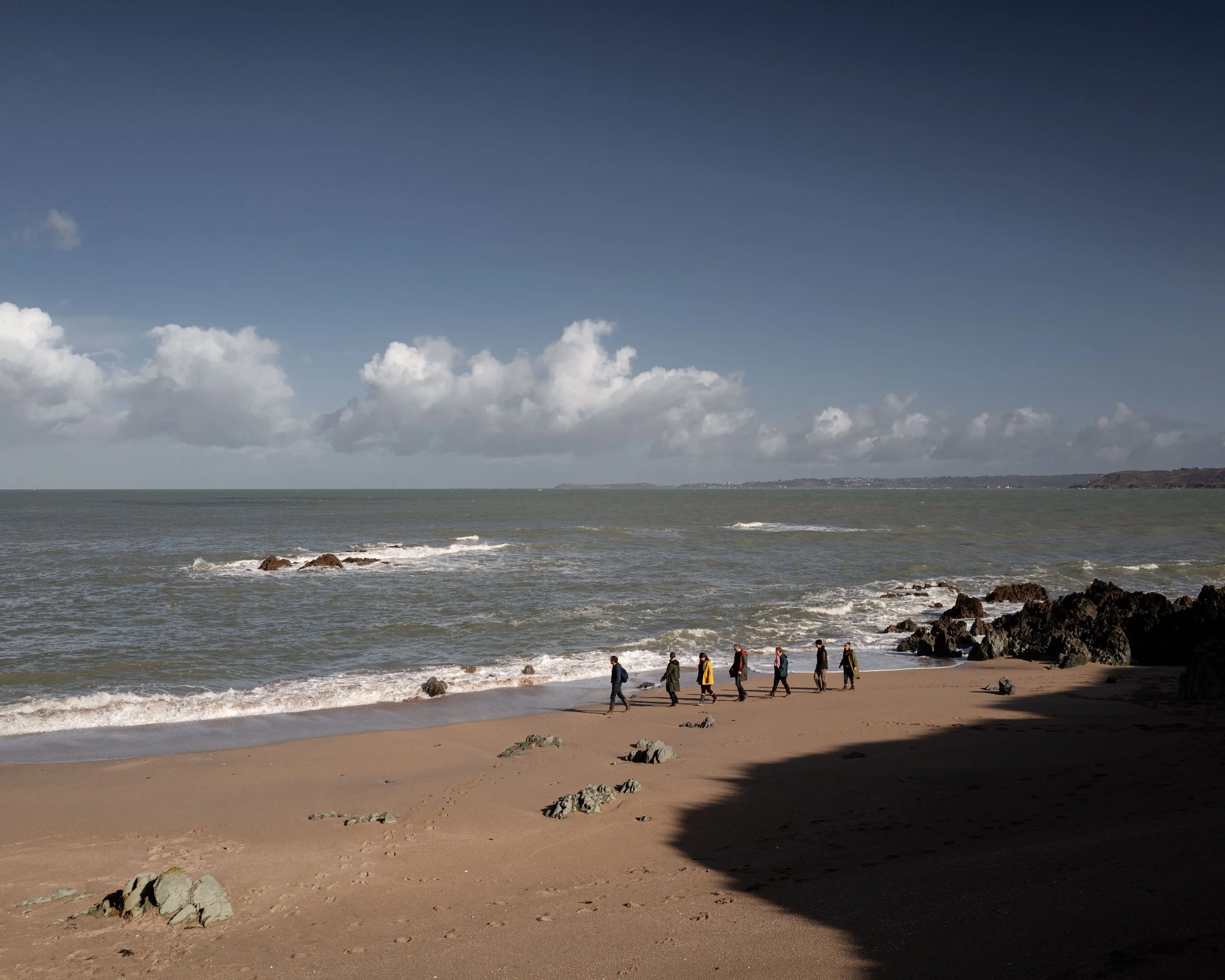 A group of people walking along a sandy beach with rocks, with the ocean and distant land in the background and a partly cloudy sky.