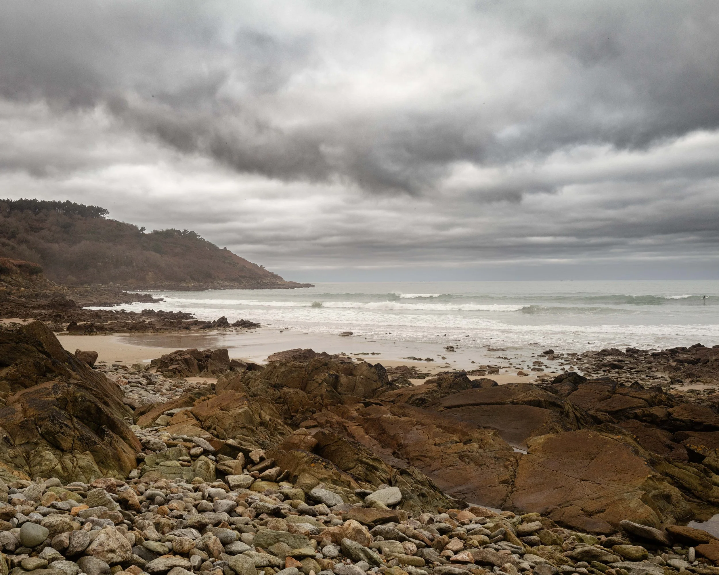 A rocky beach shoreline under a cloudy sky with waves and a distant hillside.