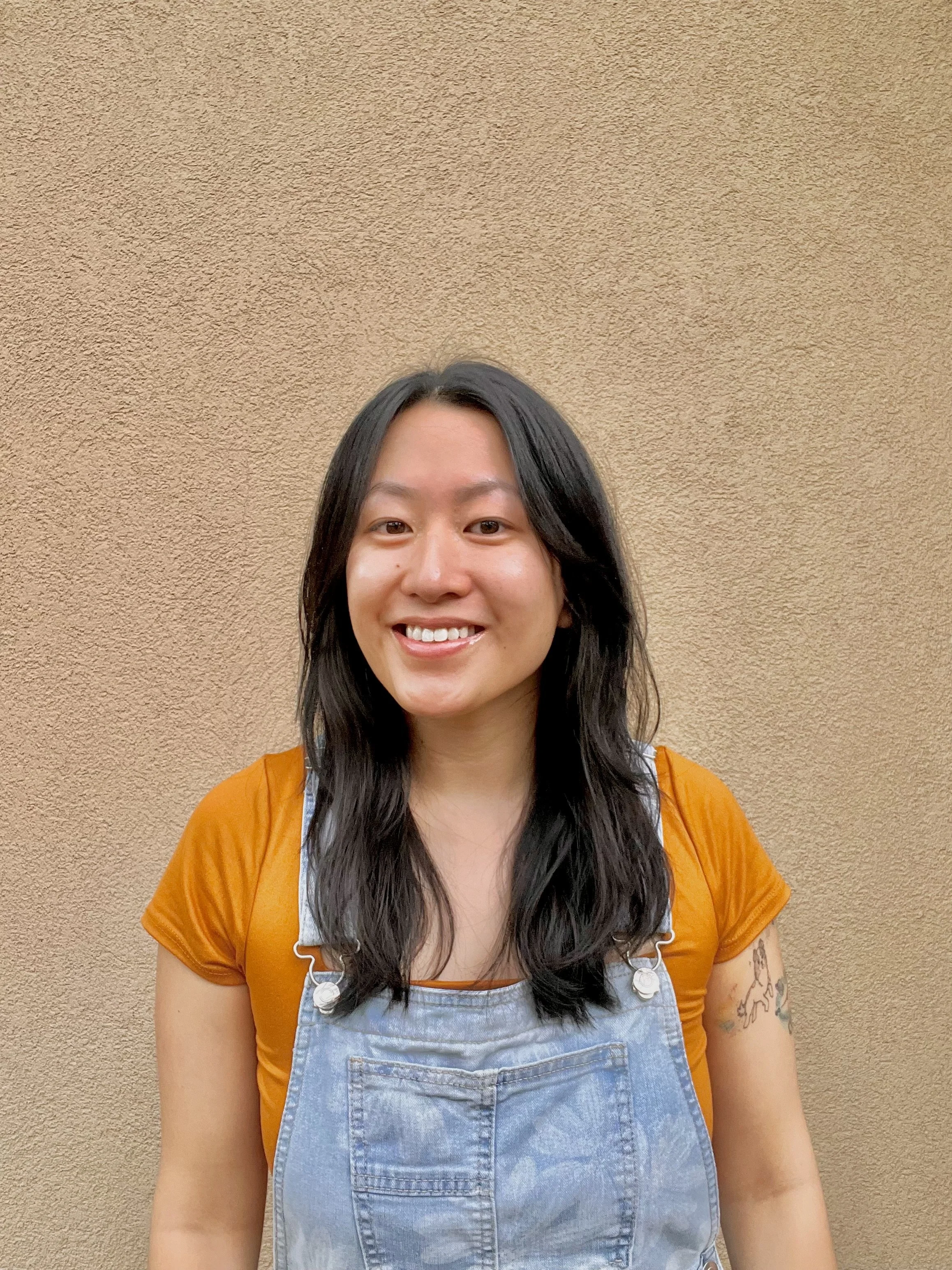 A young woman with long black hair, smiling, wearing an orange shirt and denim overalls, standing against a beige wall.