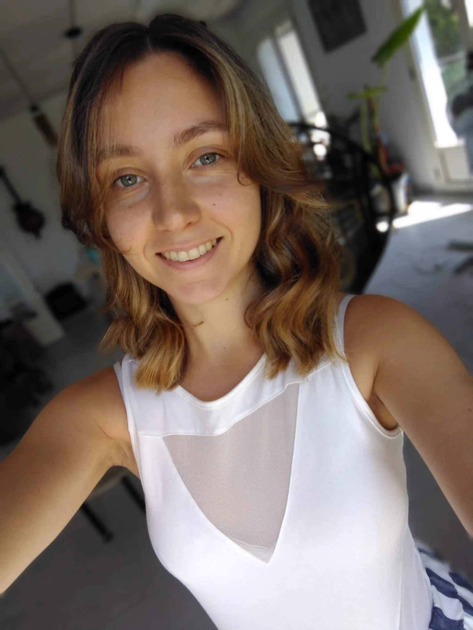 Une jeune femme souriante dans une pièce, portant un haut blanc avec des détails en mesh, avec des murs blancs, une fenêtre, et des objets en arrière-plan.