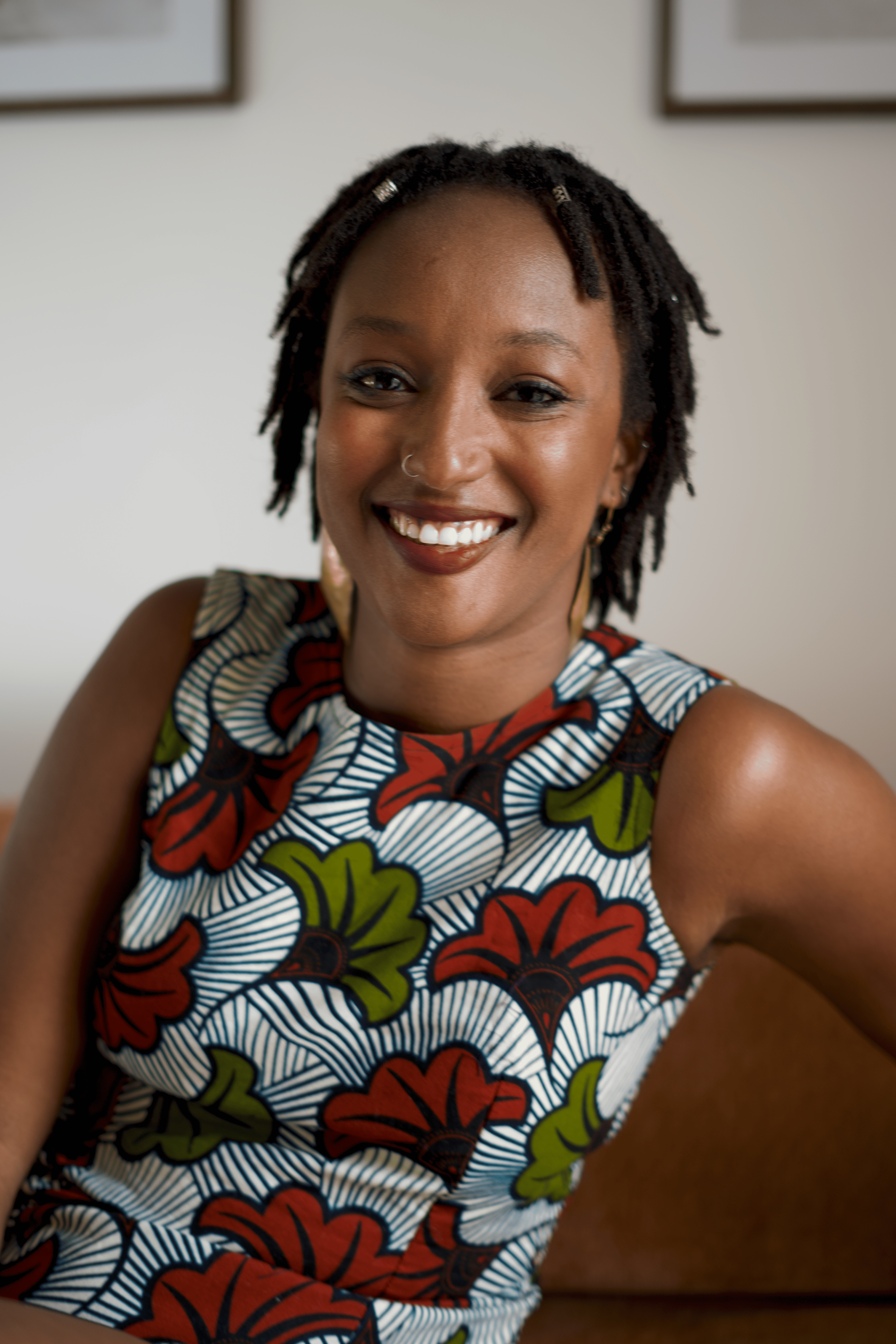 Une femme souriante, avec des dreadlocks, portant une robe colorée avec un motif floral, dans un intérieur avec des cadres au mur.