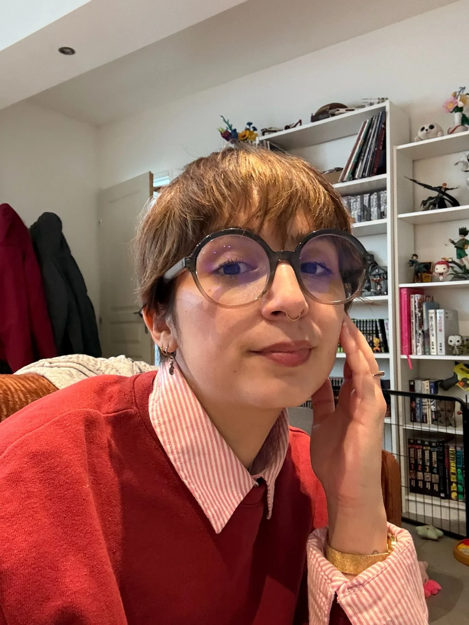 Une femme porte des lunettes rondes, des boucles d'oreilles et un collier, elle a des cheveux courts bruns et un maquillage léger. Elle est assise dans une pièce avec une étagère remplie de livres et de figurines en arrière-plan.