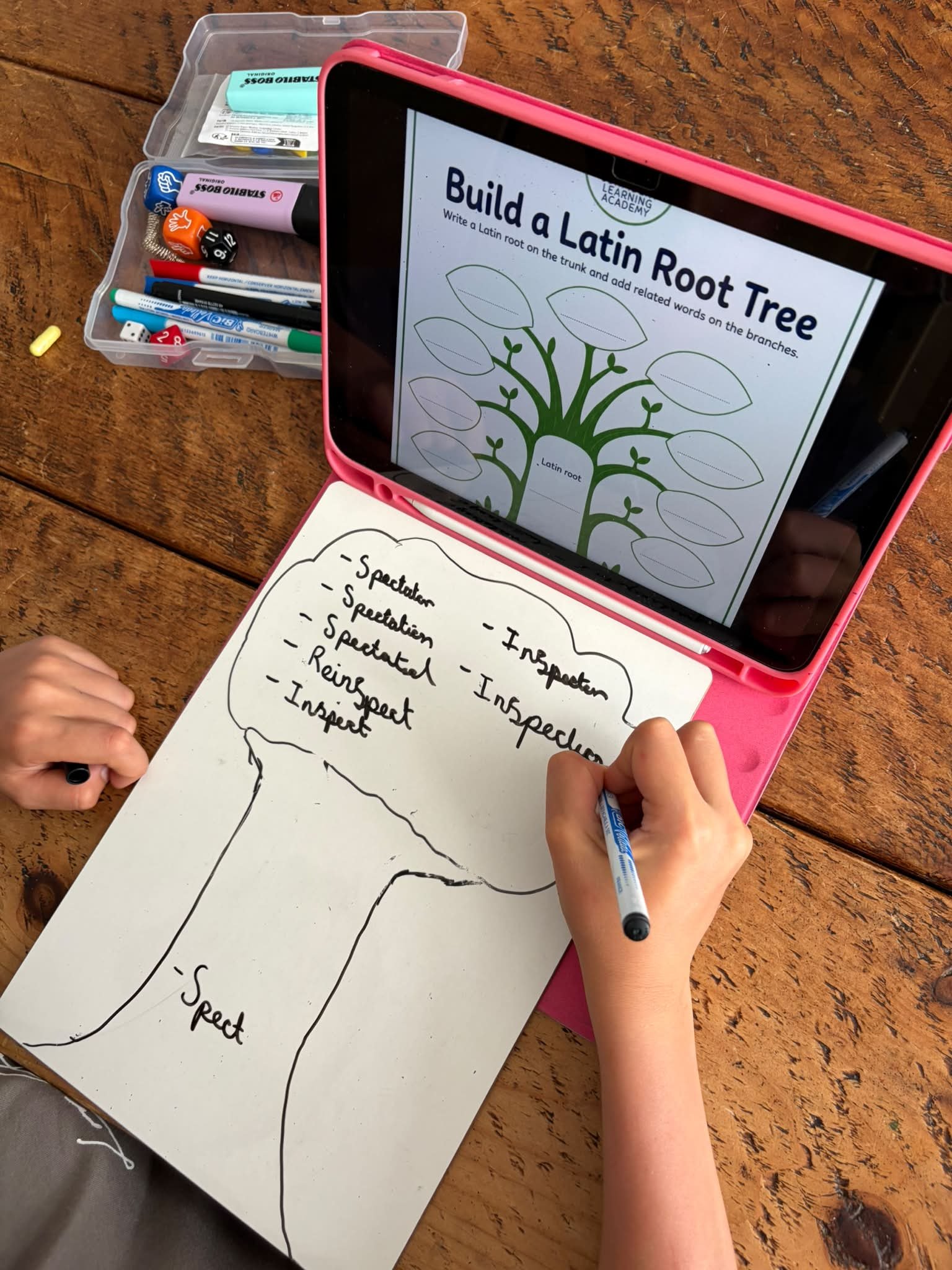 A child is drawing a diagram of a tree on a white sheet of paper. The child is wearing a bracelet on their wrist and uses a black pen. The paper has a large tree outline with blank spaces for writing. An electronic device on the table displays an educational activity titled 'Build a Latin Root Tree' with an illustration of a tree with blank leaves for Latin roots and related words. There is a plastic container with various colored markers and a yellow chalk on the wooden table.
