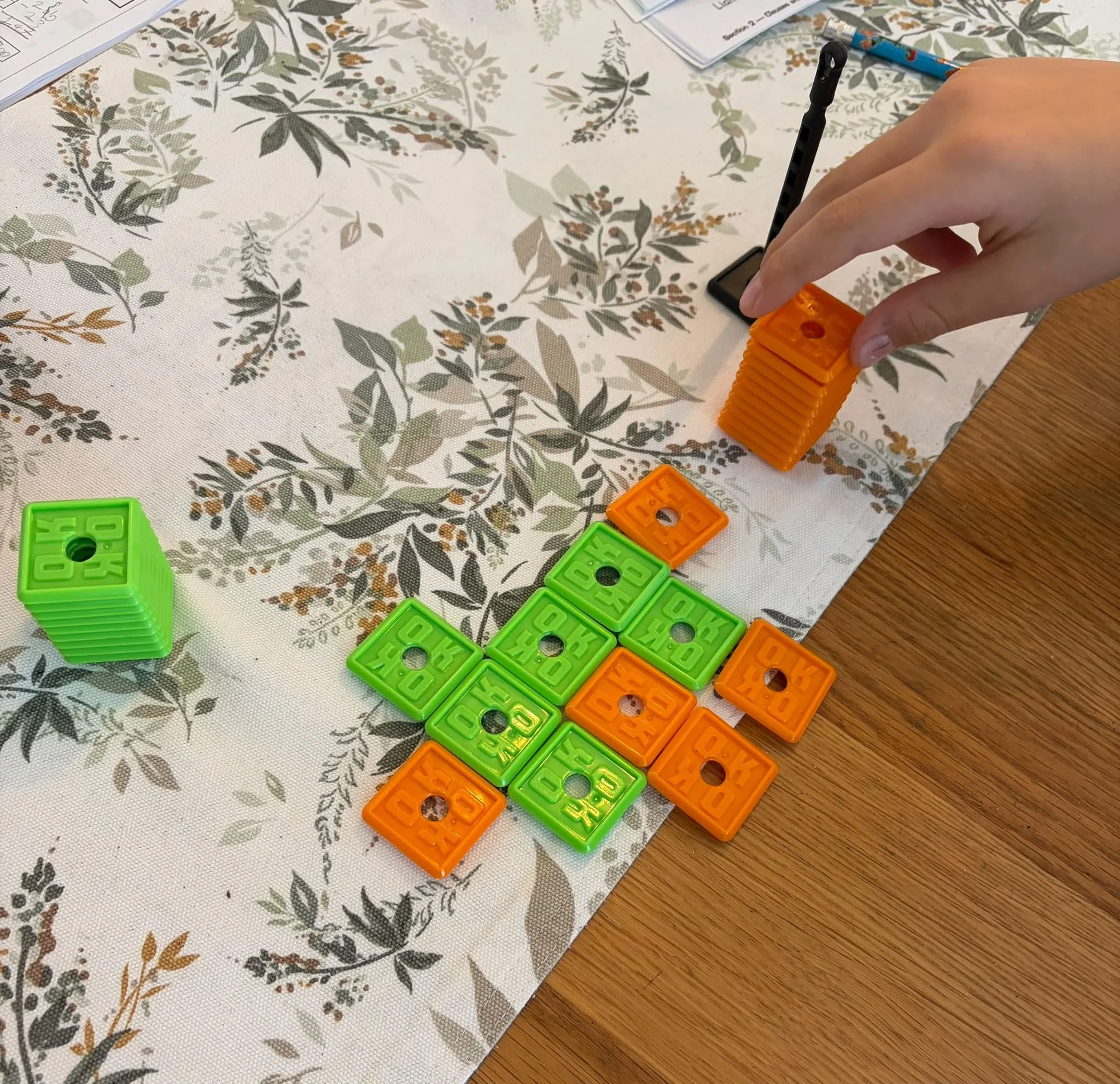A person's hand is holding an orange game piece on a tabletop covered with a patterned cloth. There are several green and orange game pieces scattered on the table, which are rectangular with holes and the word 'blocks' embossed. The scene suggests a board game or puzzle activity.