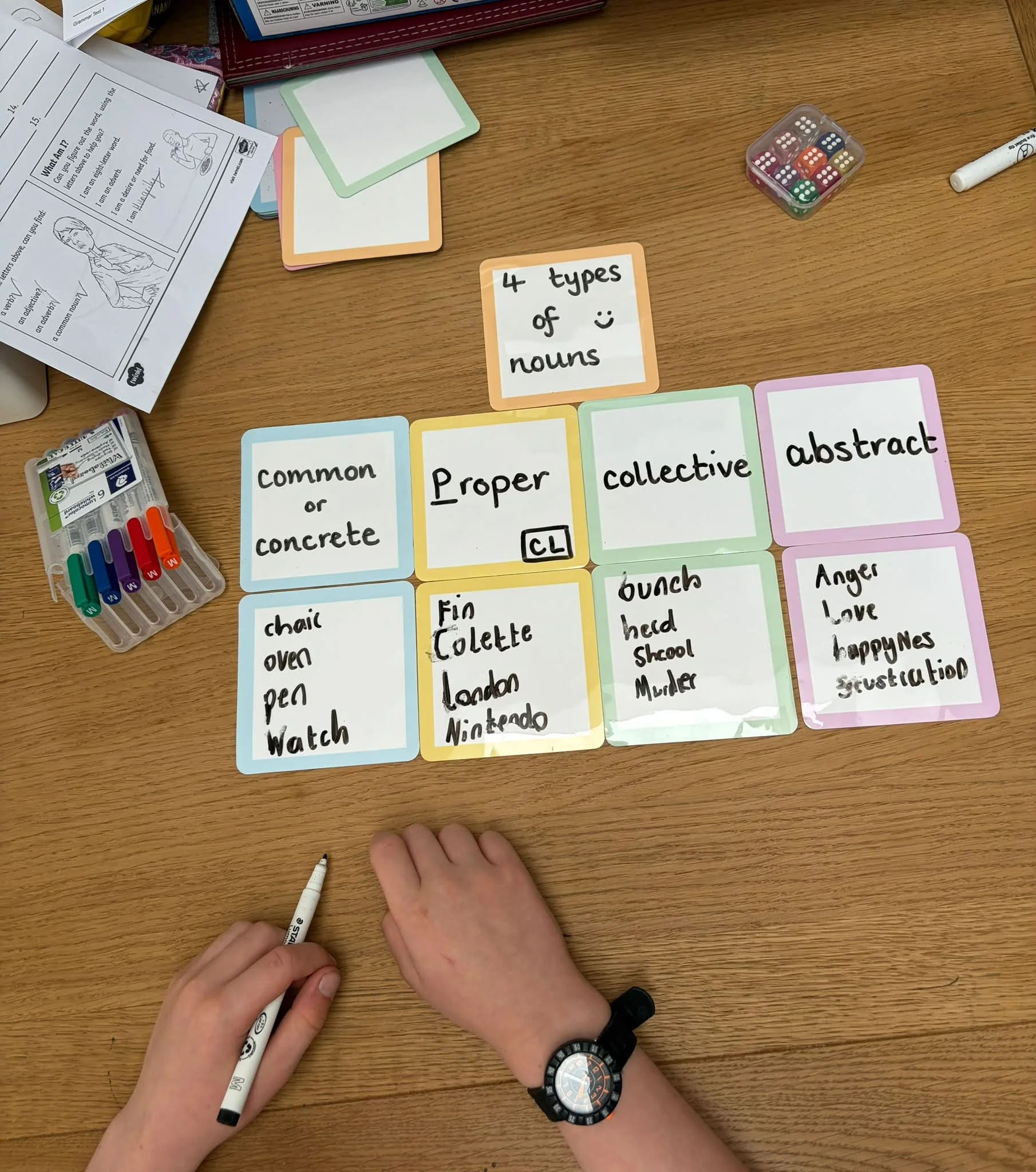 Hands of a child writing on flashcards with a black marker pen, with colorful flashcards laid out on a wooden table and a container of multicolored dice nearby.