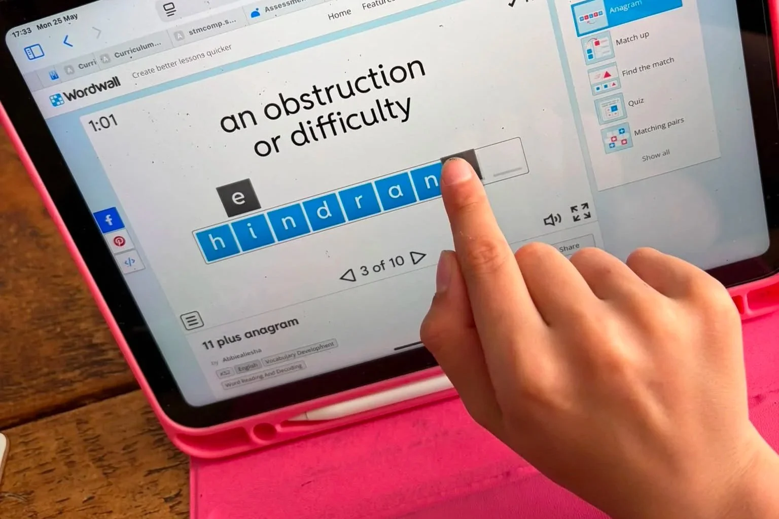 A person using a tablet with a pink case to solve a word puzzle on a language learning app. The puzzle shows the prefix 'e' and the partially completed word 'hindran' with spaces for the missing letters, under the hint 'an obstruction or difficulty.'