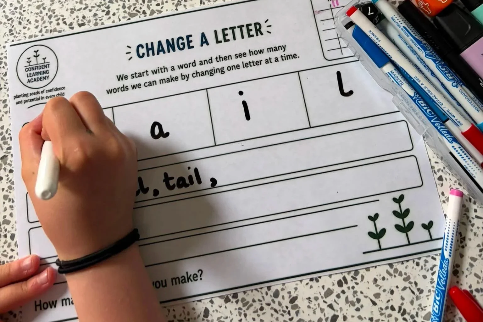 Children's worksheet titled 'Change a Letter' for phonics practice, with handwritten words 'a' and 'i', and a partially written word 'tail,' surrounded by various colored markers and pens on a speckled tabletop.