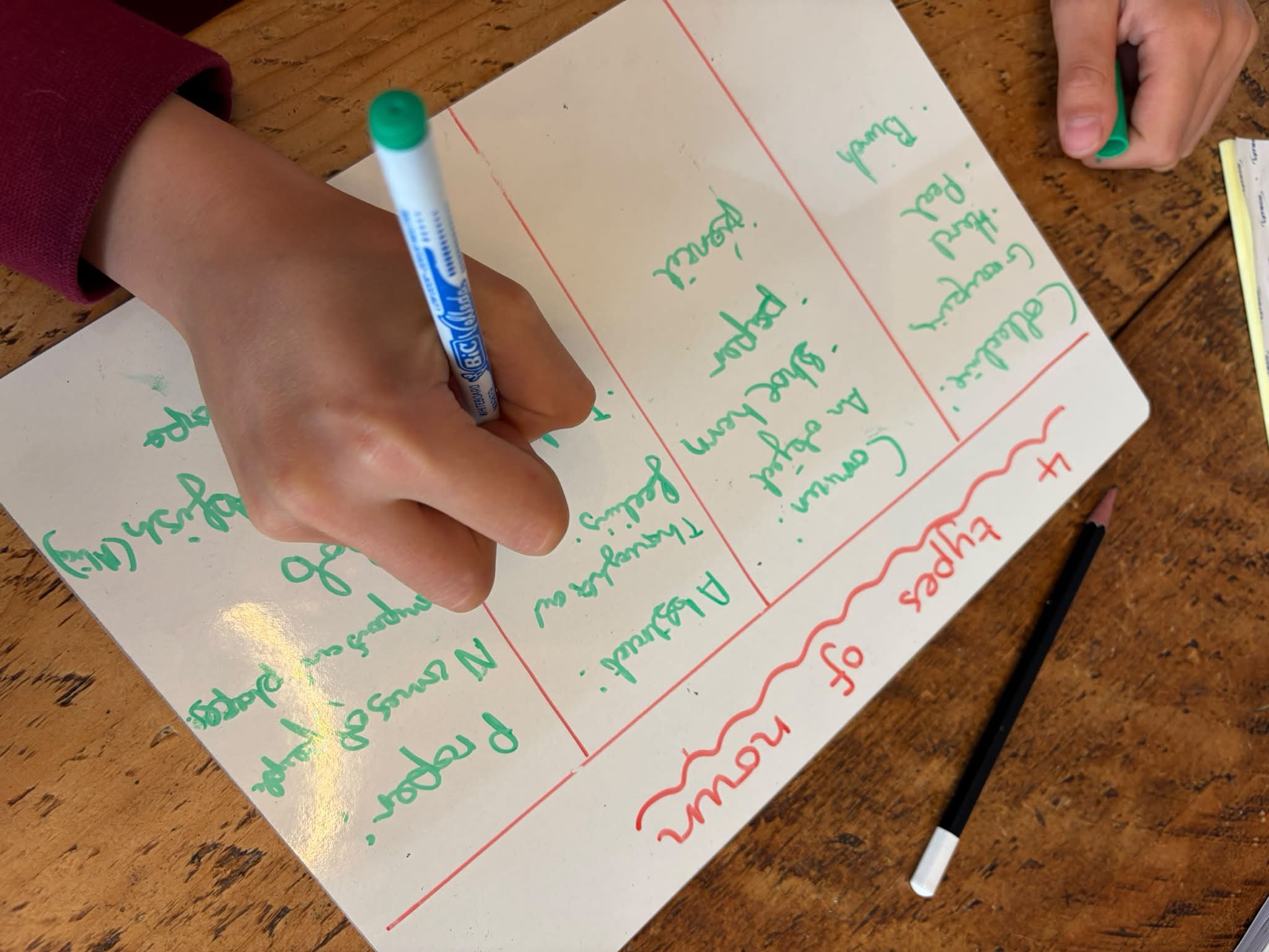 A child writes on a white paper with green and red markers, with a piece of paper and a black marker on a wooden table.