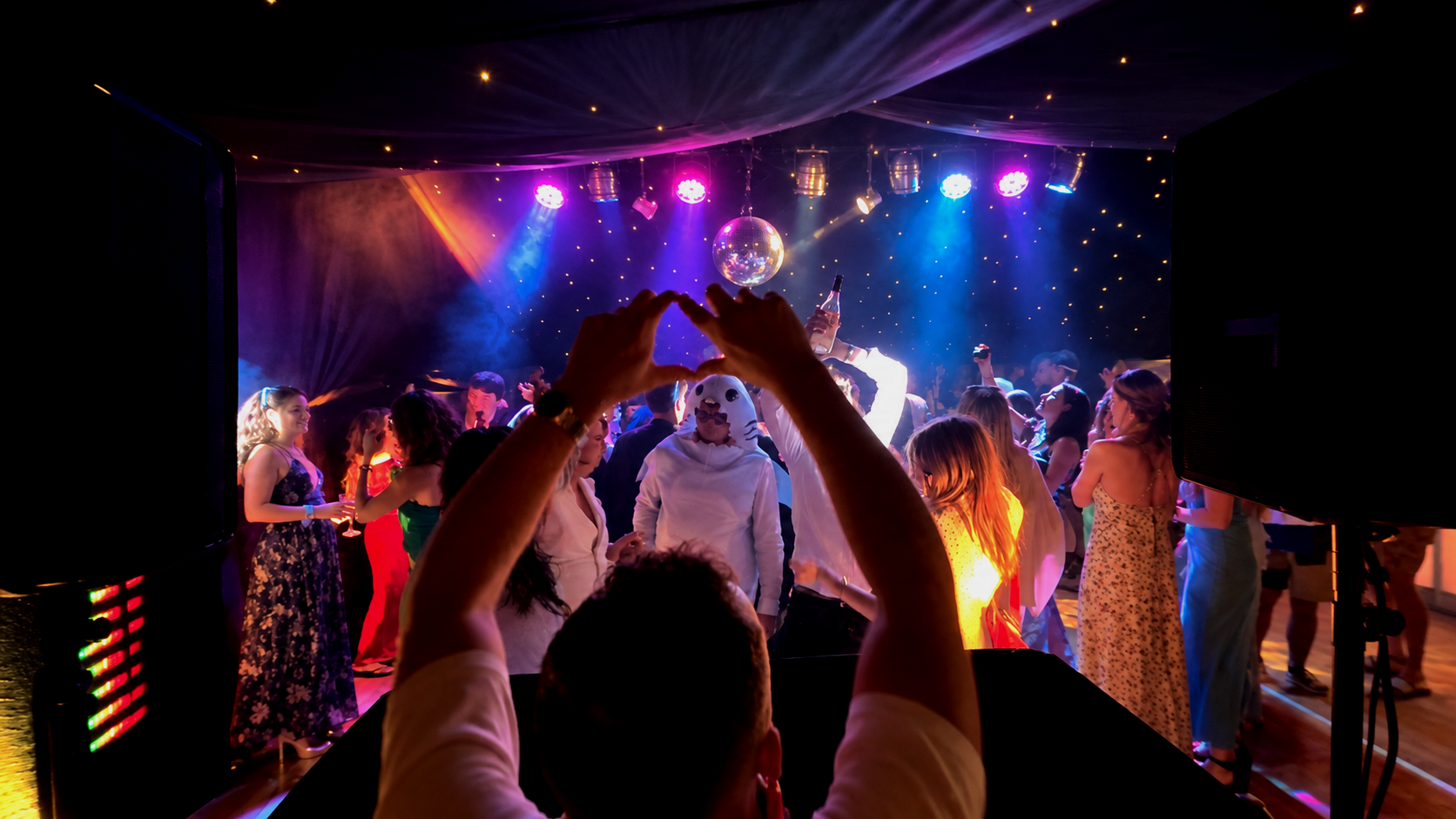 People dancing and celebrating at a nightclub with colorful lights, a disco ball, and a person wearing a dog mask.