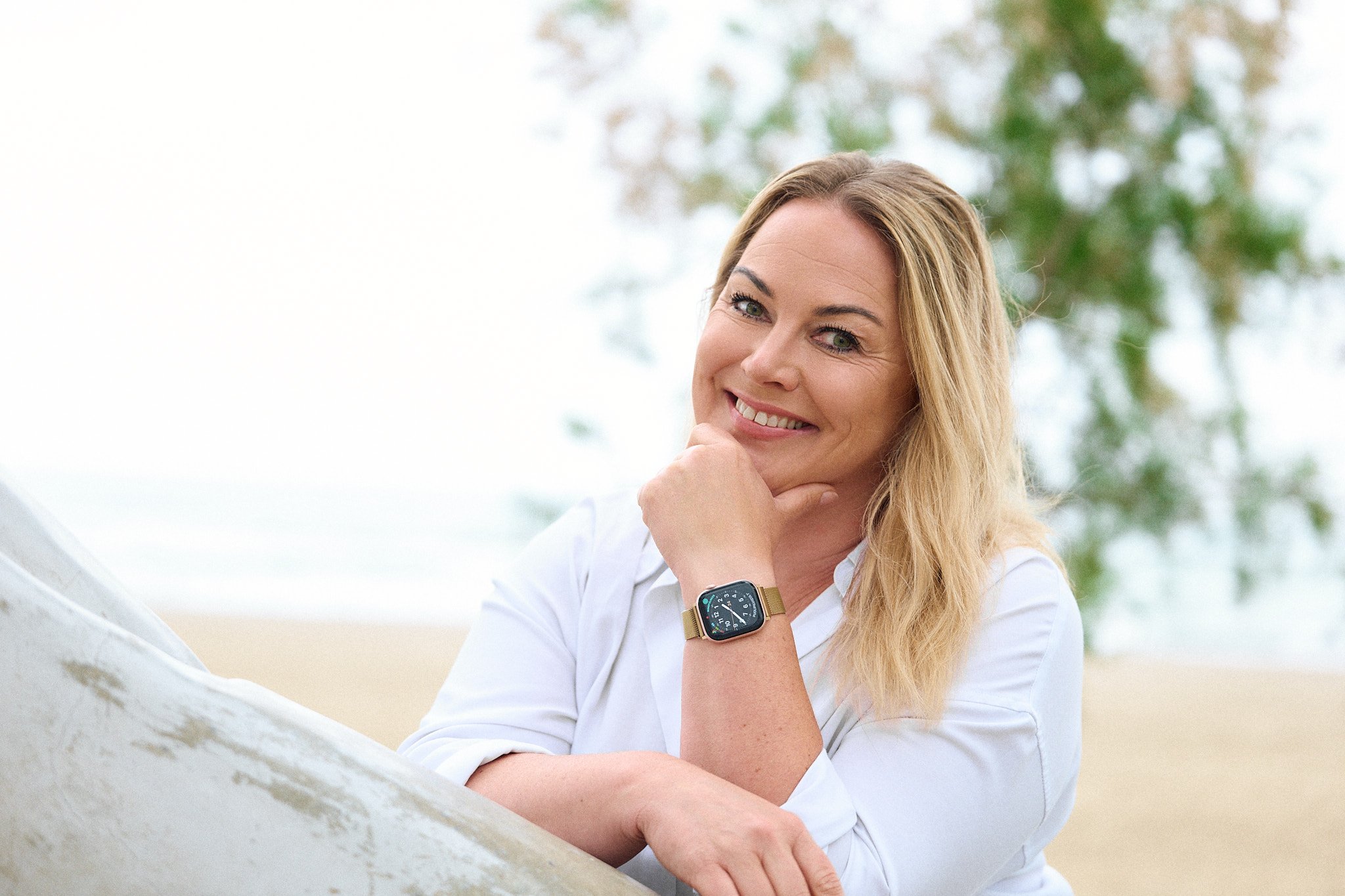 A woman with blonde hair and green eyes, smiling and resting her chin on her hand, wearing a smartwatch and a white shirt outdoors with trees and sky in the background.