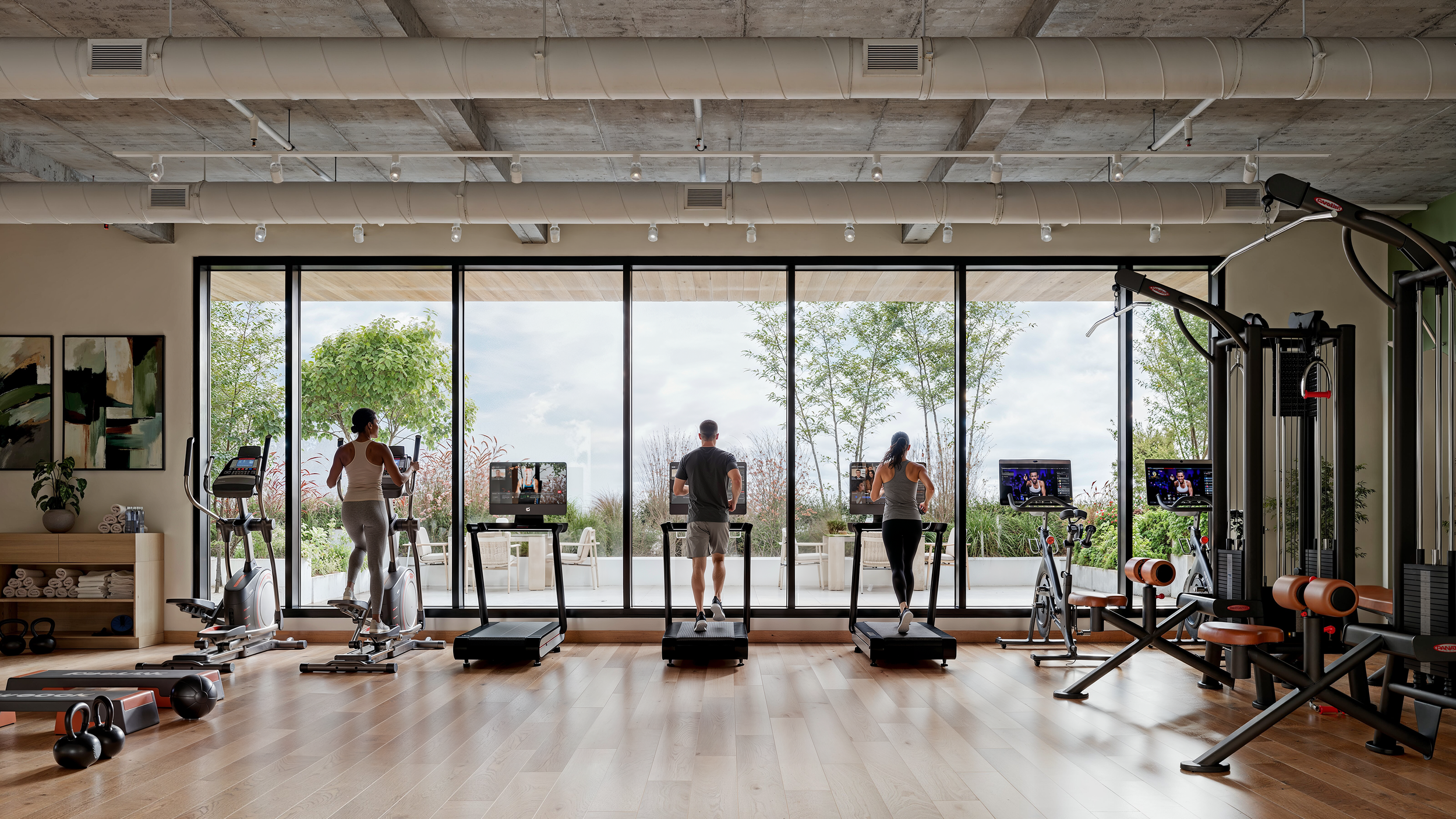 Indoor gym with three people using cardio machines, large window overlooking outdoor greenery, and various gym equipment.