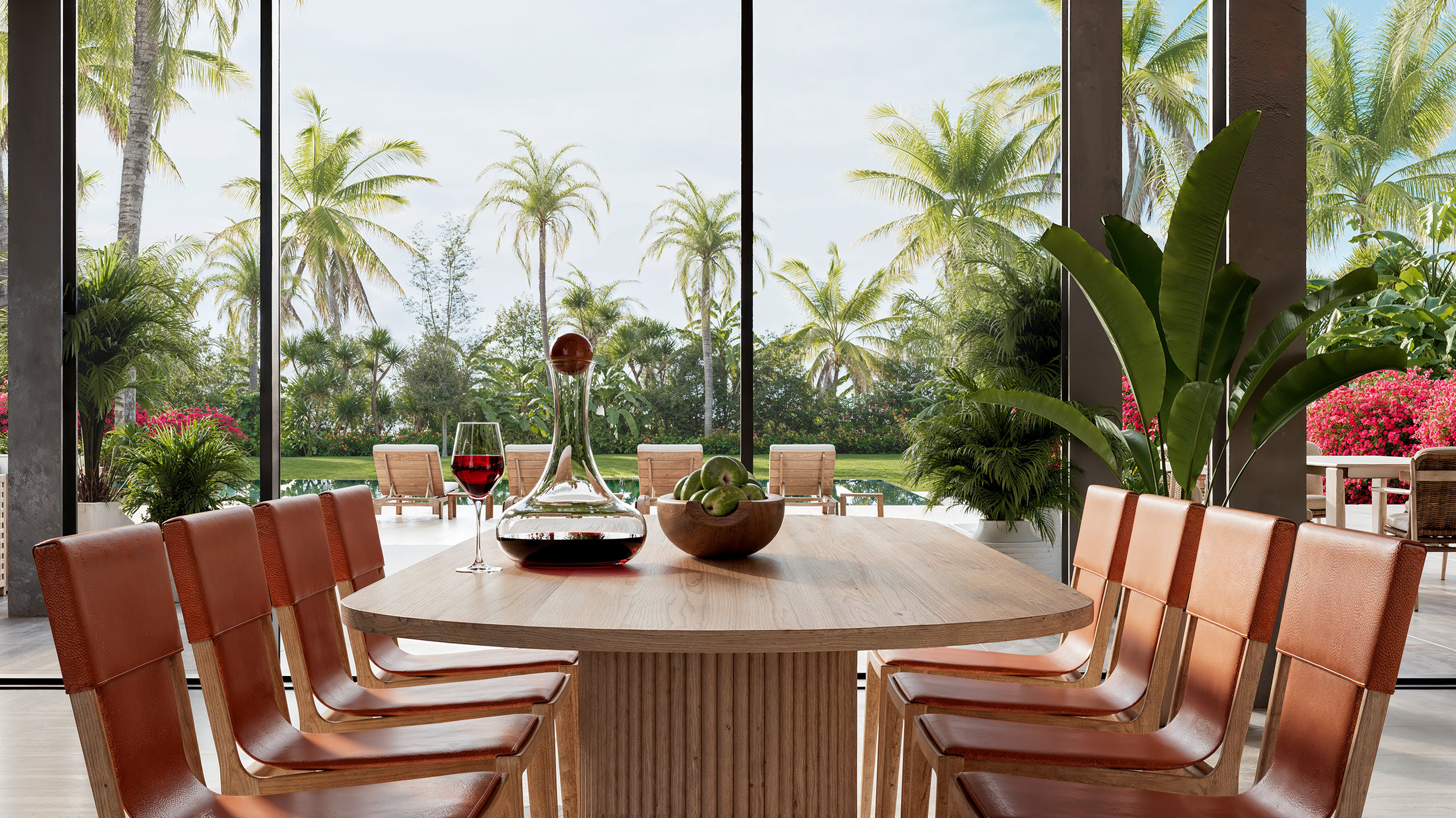 Dining room with a wooden table, chairs, and a view of a lush tropical garden with palm trees through large glass doors.