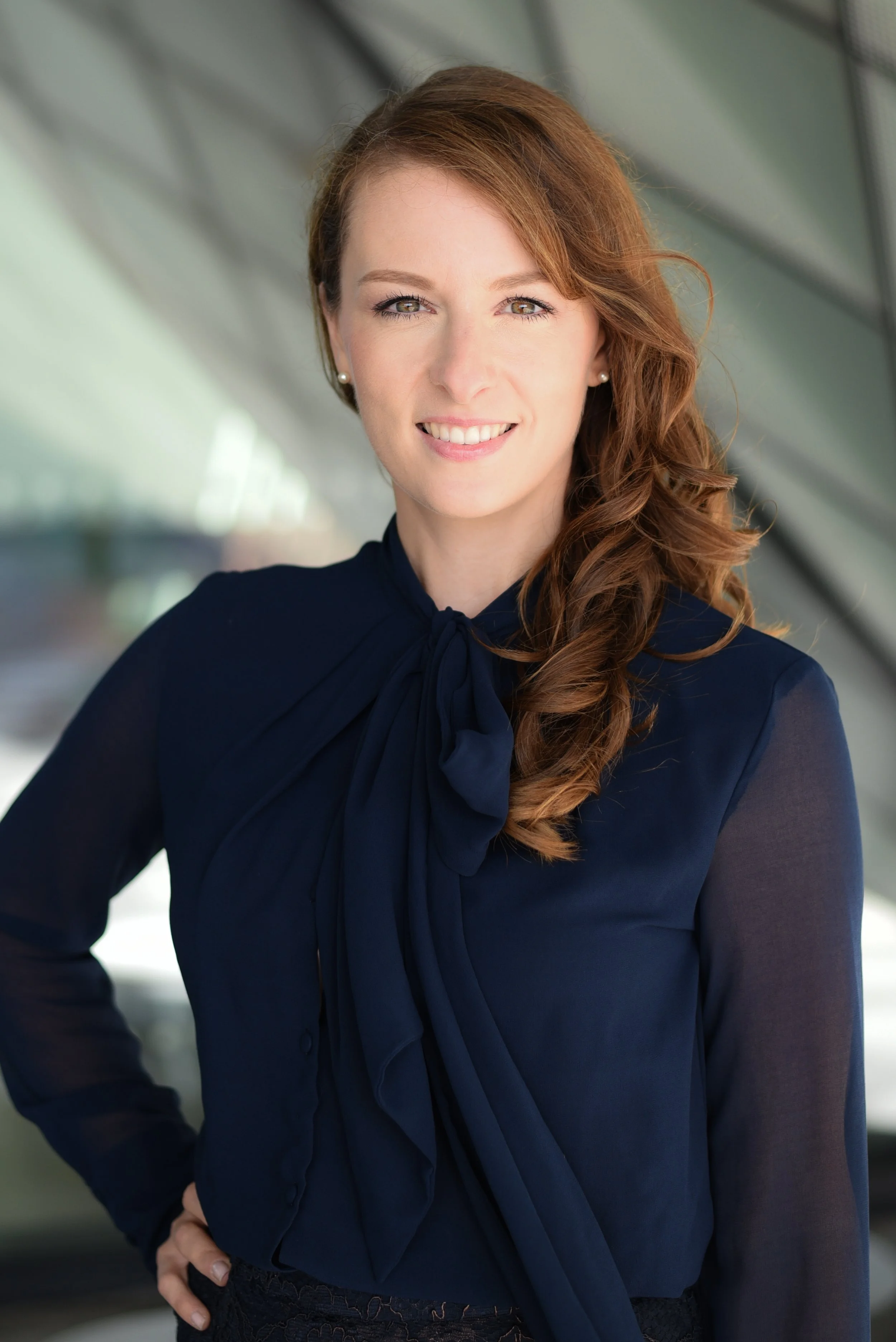 Veronika Westerheide, mit langem, lockigem, rotbraunem Haar trägt eine dunkelblaue Bluse mit Schleife am Hals und perlenschmuck. Sie steht vor einem modernen, glasüberdachten Gebäude, dem Futurium Berlin, und lächelt in die Kamera.