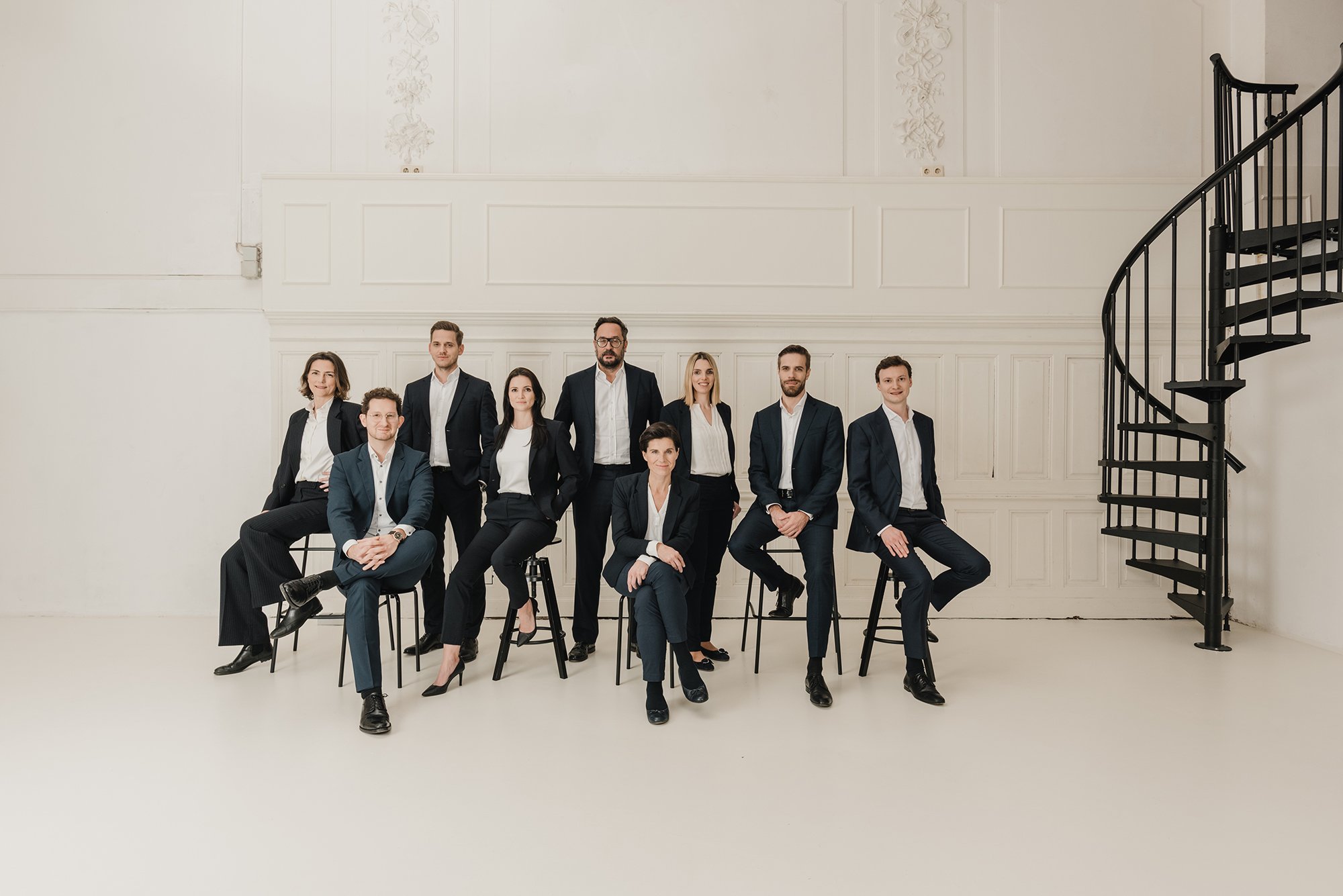 Eine Gruppe von zehn Menschen in formeller Kleidung posiert für ein Gruppenfoto in einem hellen, eleganten Raum mit weißen Wänden und einer schwarzen Wendeltreppe.