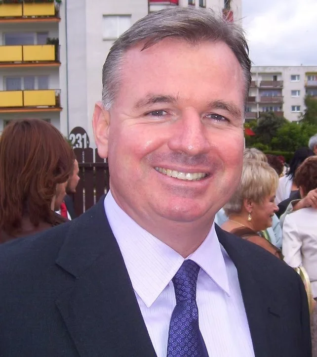 Smiling man in a suit with a white shirt and purple tie at an outdoor event, with people and residential buildings in the background.