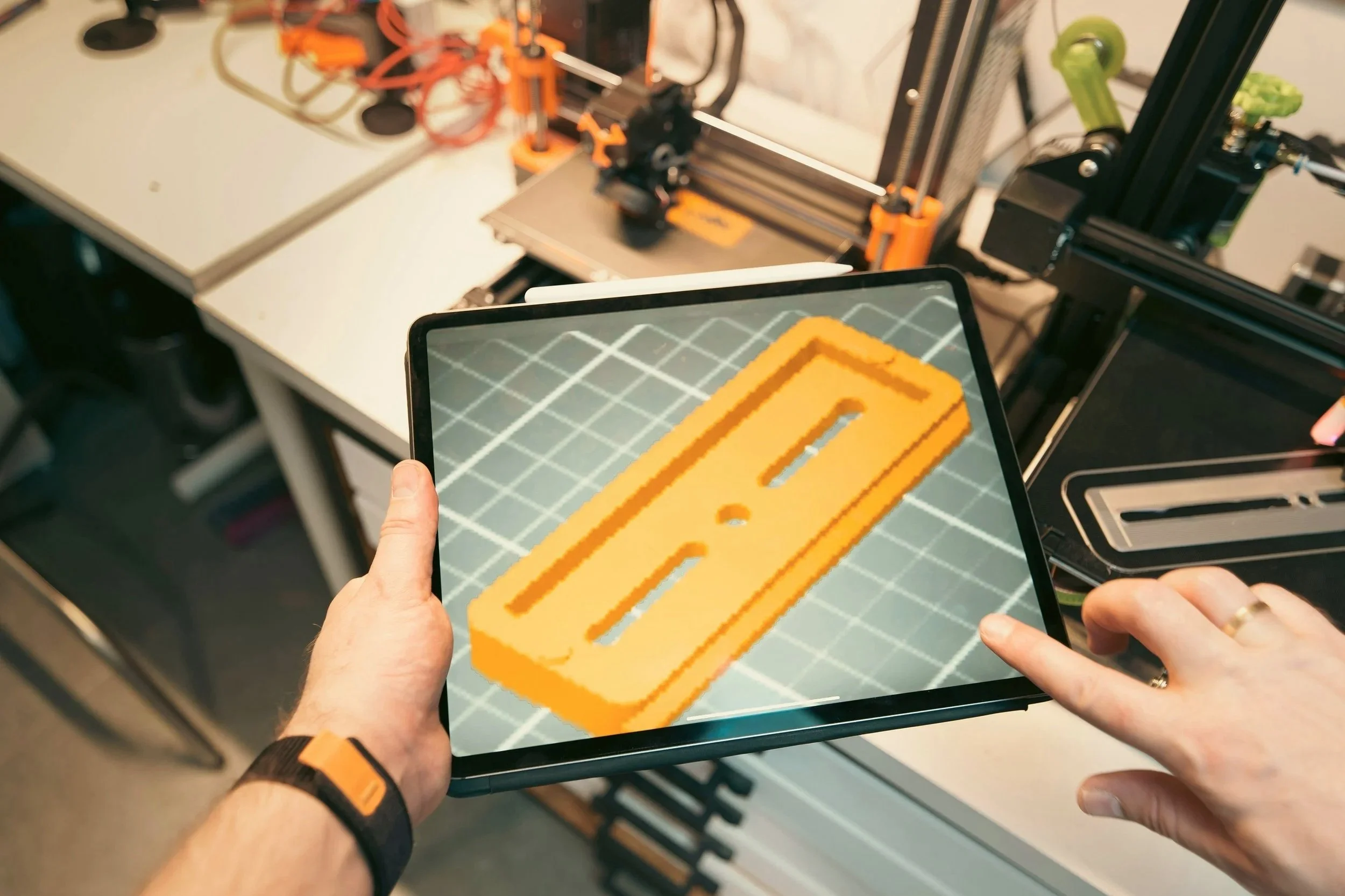 Person holding a tablet displaying a digital design of an orange and yellow pixelated object, possibly a 3D printer in the background.
