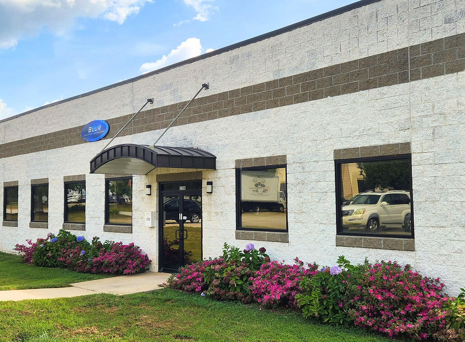 Exterior view of a commercial building with a sign that reads 'BLUR Machine Laboratory'; building features white brick walls with dark brown accents and flower beds with pink and purple hydrangeas in front.