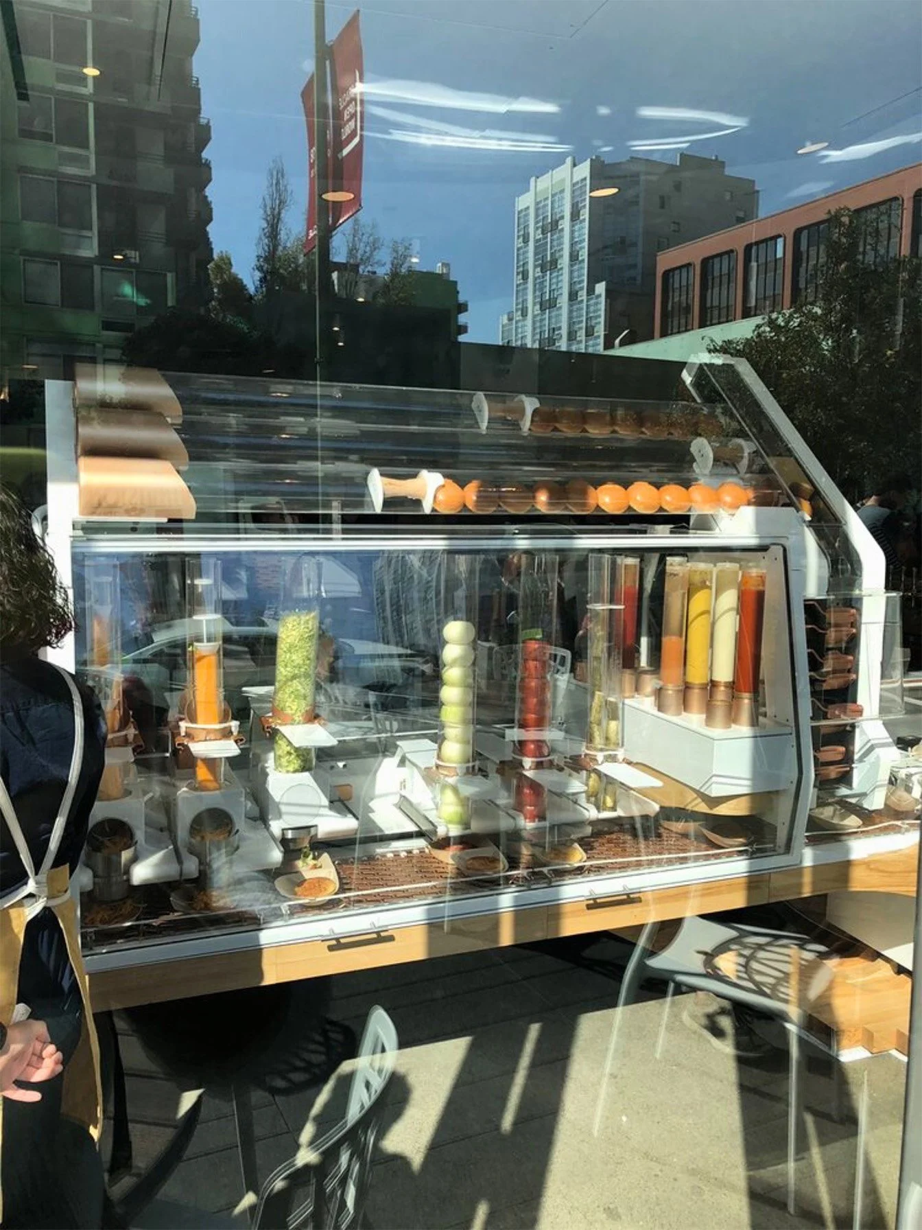 Display case filled with various frozen desserts and toppings, with reflections of buildings and a cloudy sky in the glass window.