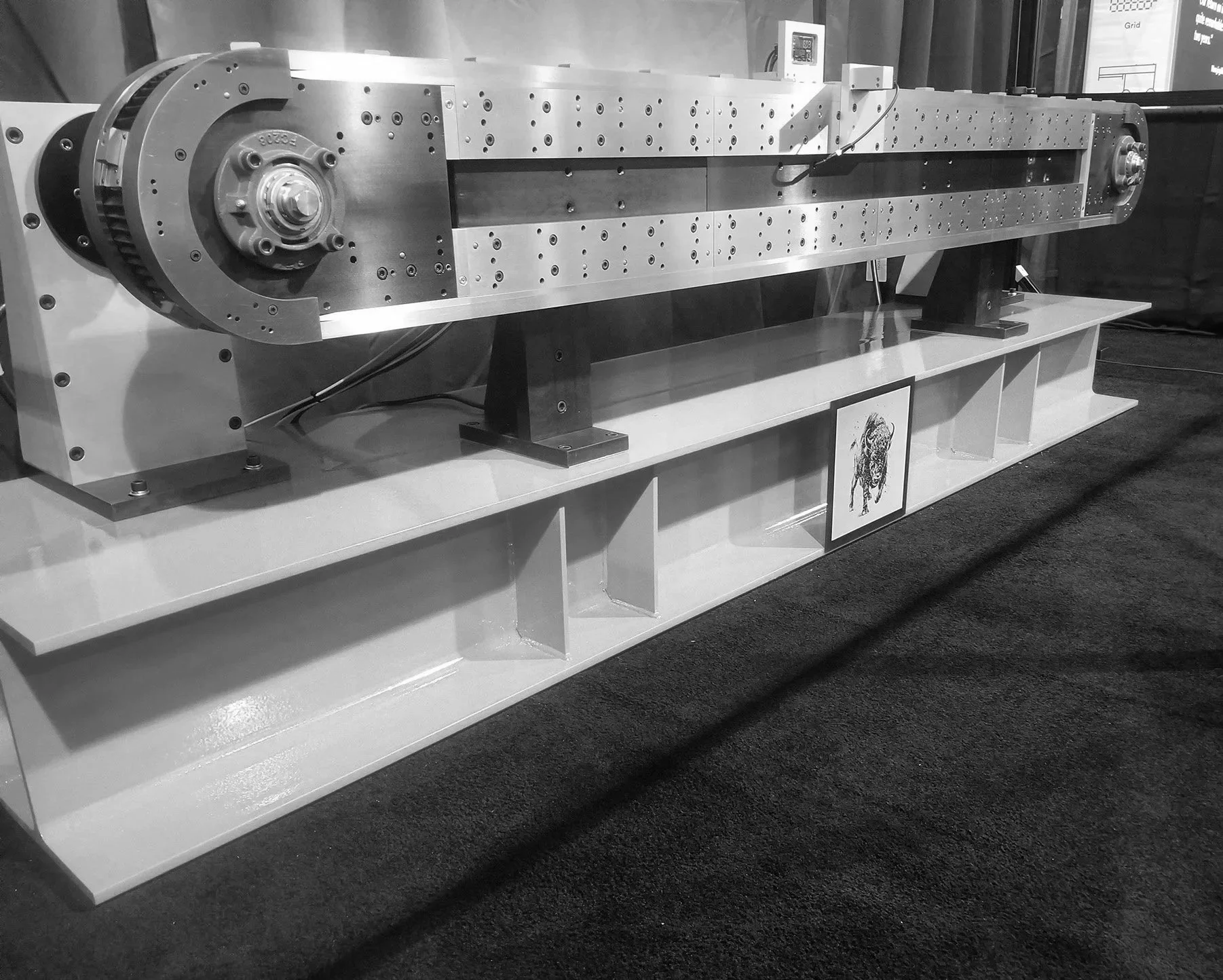A large metallic mechanical device, possibly a linear actuator or a part of industrial machinery, displayed on a platform.