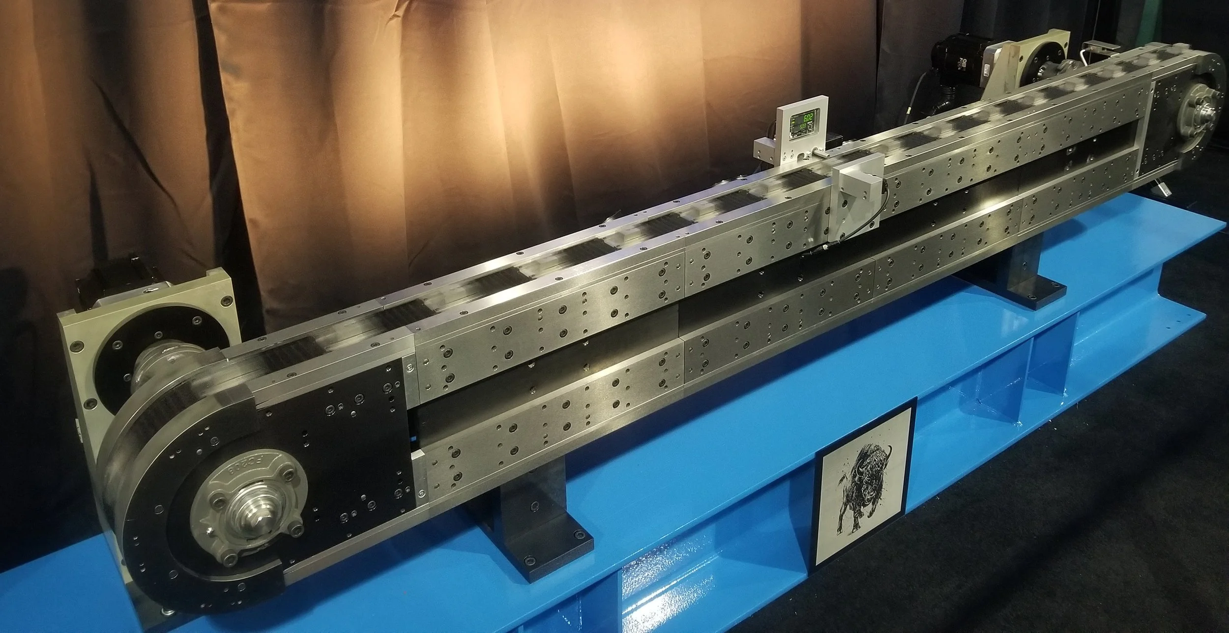 A long metallic device with rollers, sensors, and electronic components mounted on a blue display stand, with a black curtain background.