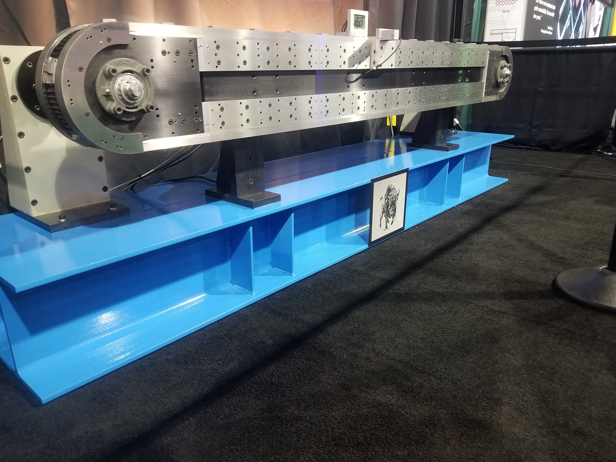 A large, elongated metal industrial machine with circular components on each end, mounted on a blue platform, with a black background and some informational posters on the wall behind it.