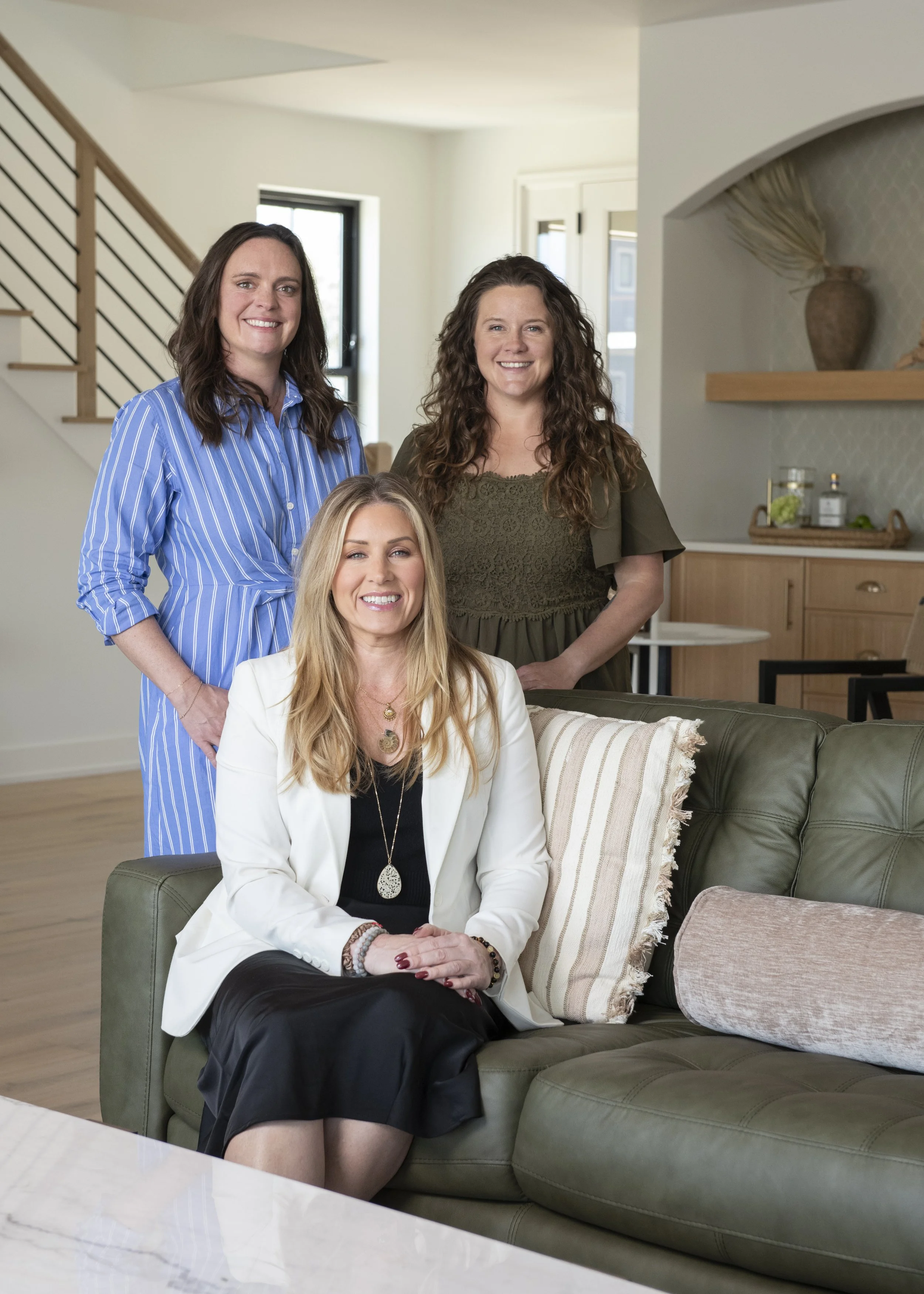 Three women in a modern living room, one sitting on a green sofa and two standing behind, all smiling at the camera.