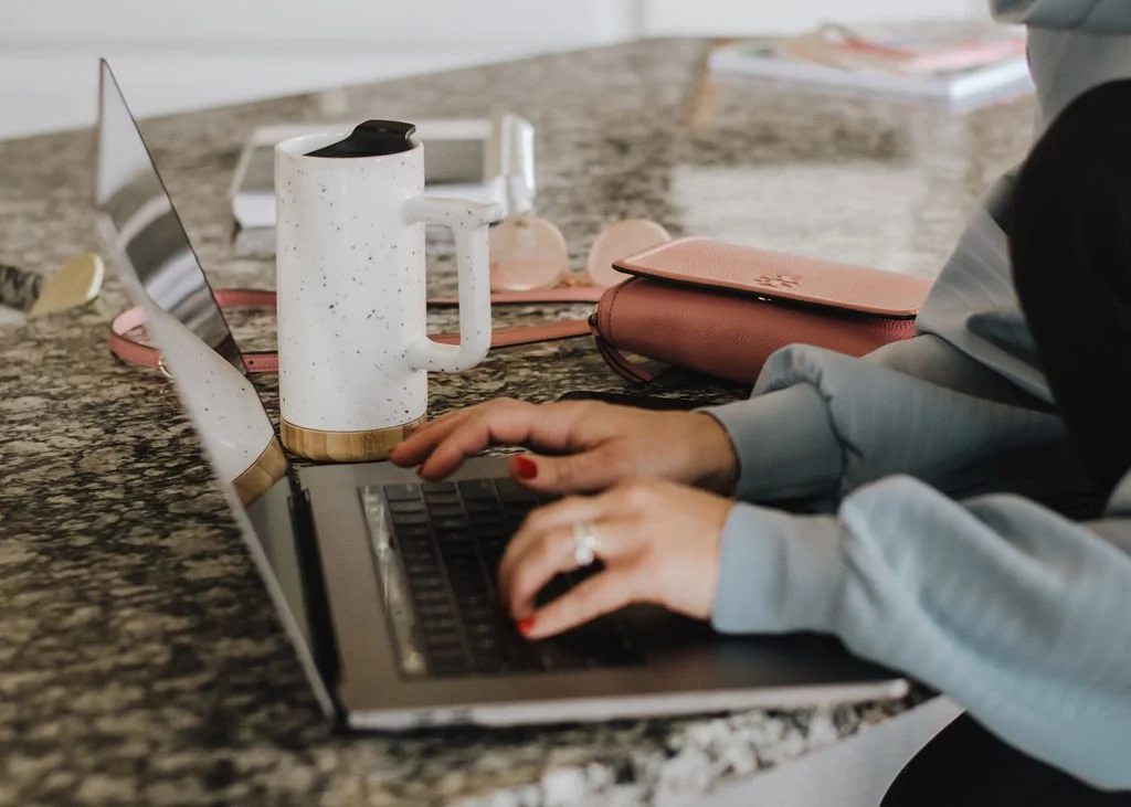 Female entrepreneur working on a laptop at a granite countertop with a coffee mug, pink clutch, and sunglasses.