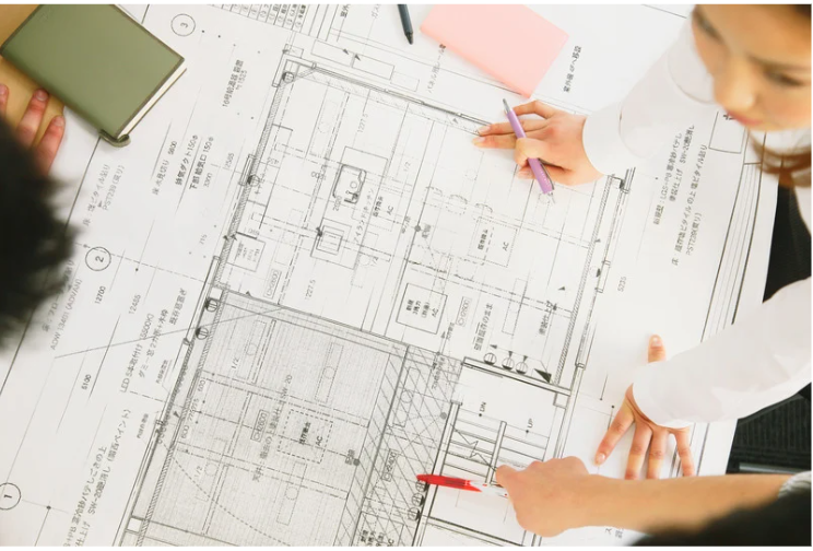 People reviewing architectural blueprints on a large table with pens, paper, and a notebook.