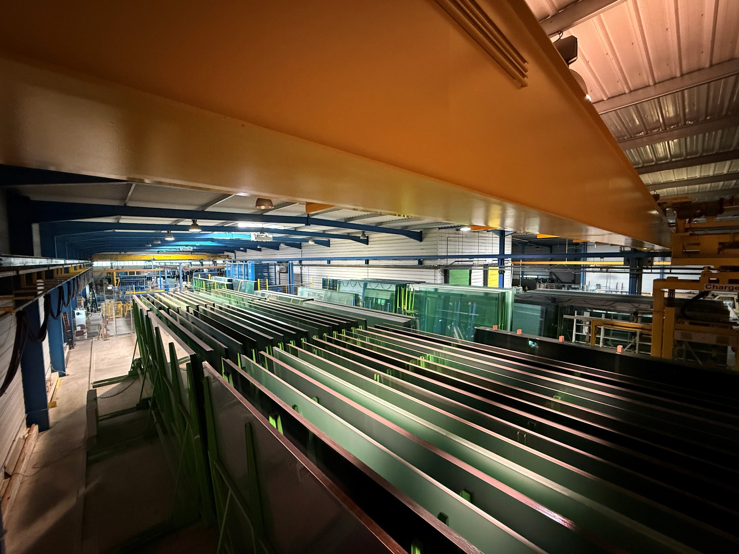 Espace de stockage de grandes plaques de verre, contenant des rangées de cadres en métal vert, des structures en acier et des panneaux en verre, dans un environnement industriel bien éclairé