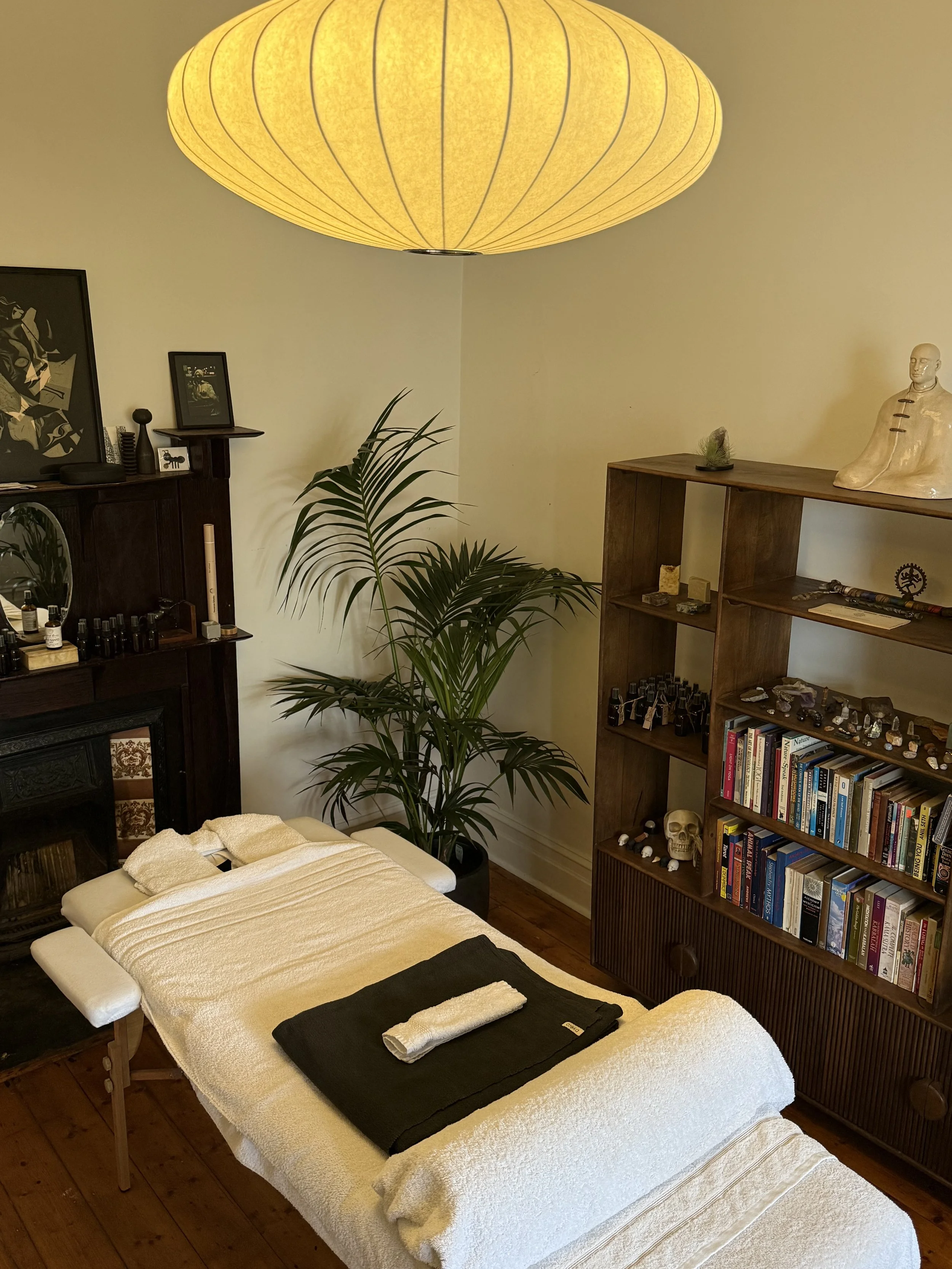 A massage table covered with folded towels in a cozy room with bookshelves, artwork, a statue, and a yellow paper lantern ceiling light.
