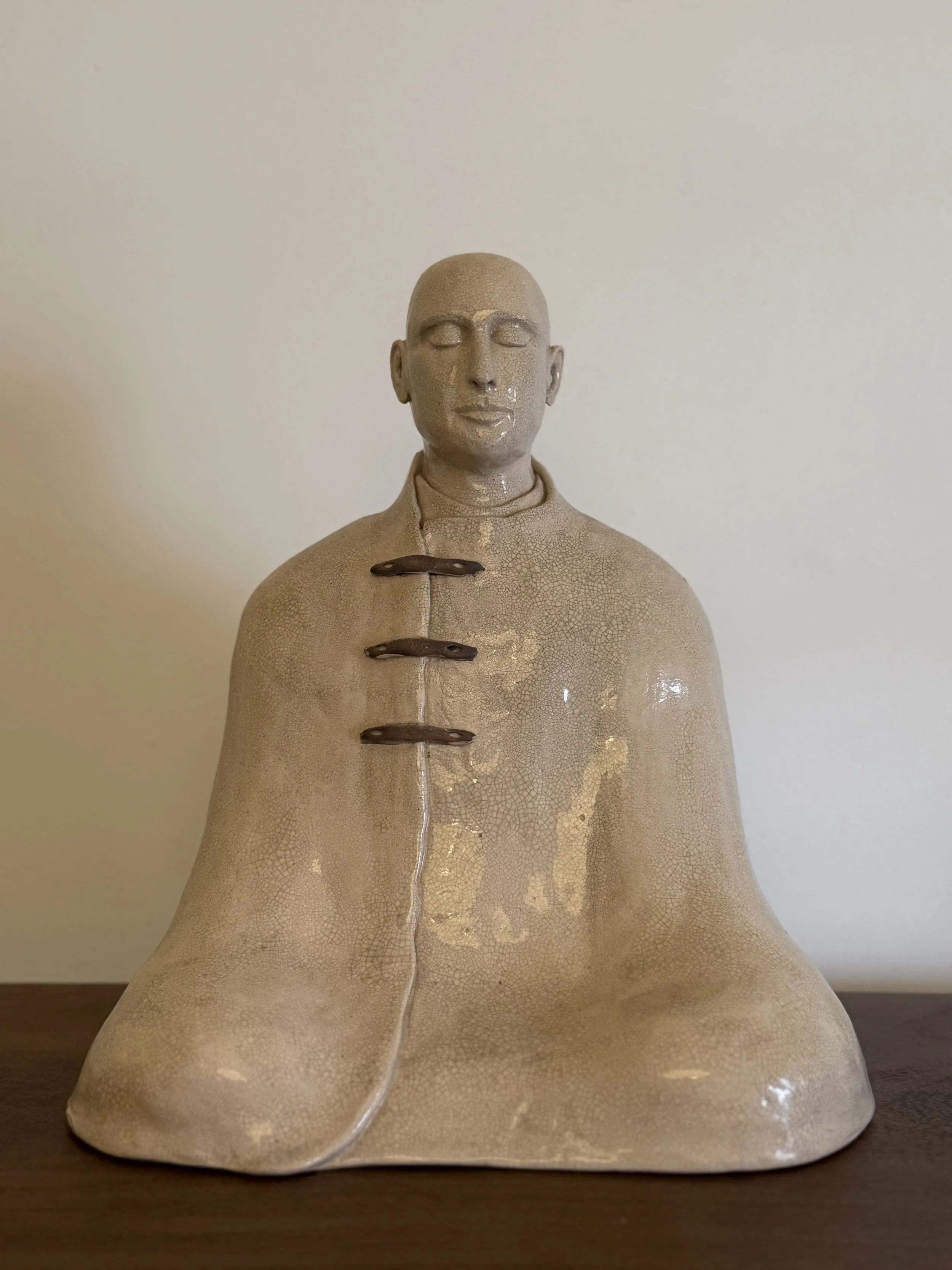Ceramic sculpture of a human figure meditating with closed eyes and a calm expression, wearing a high-collared garment with decorative clasps, set against a plain neutral background.