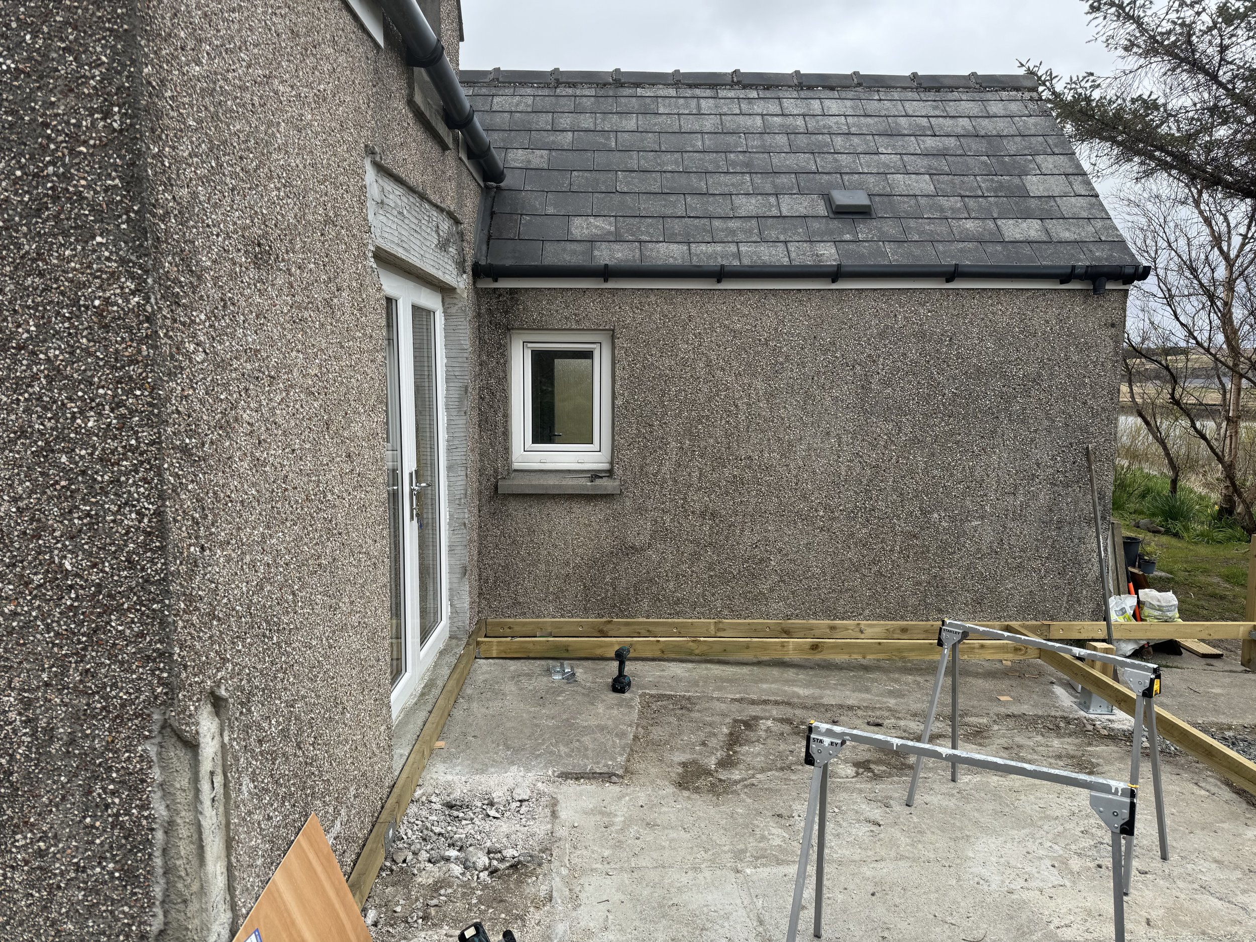 A house exterior under construction with textured grass concrete walls, a dark grey shingle roof, and a small window. The construction site has tools like a saw, cordless drill, and wooden frame on the ground.