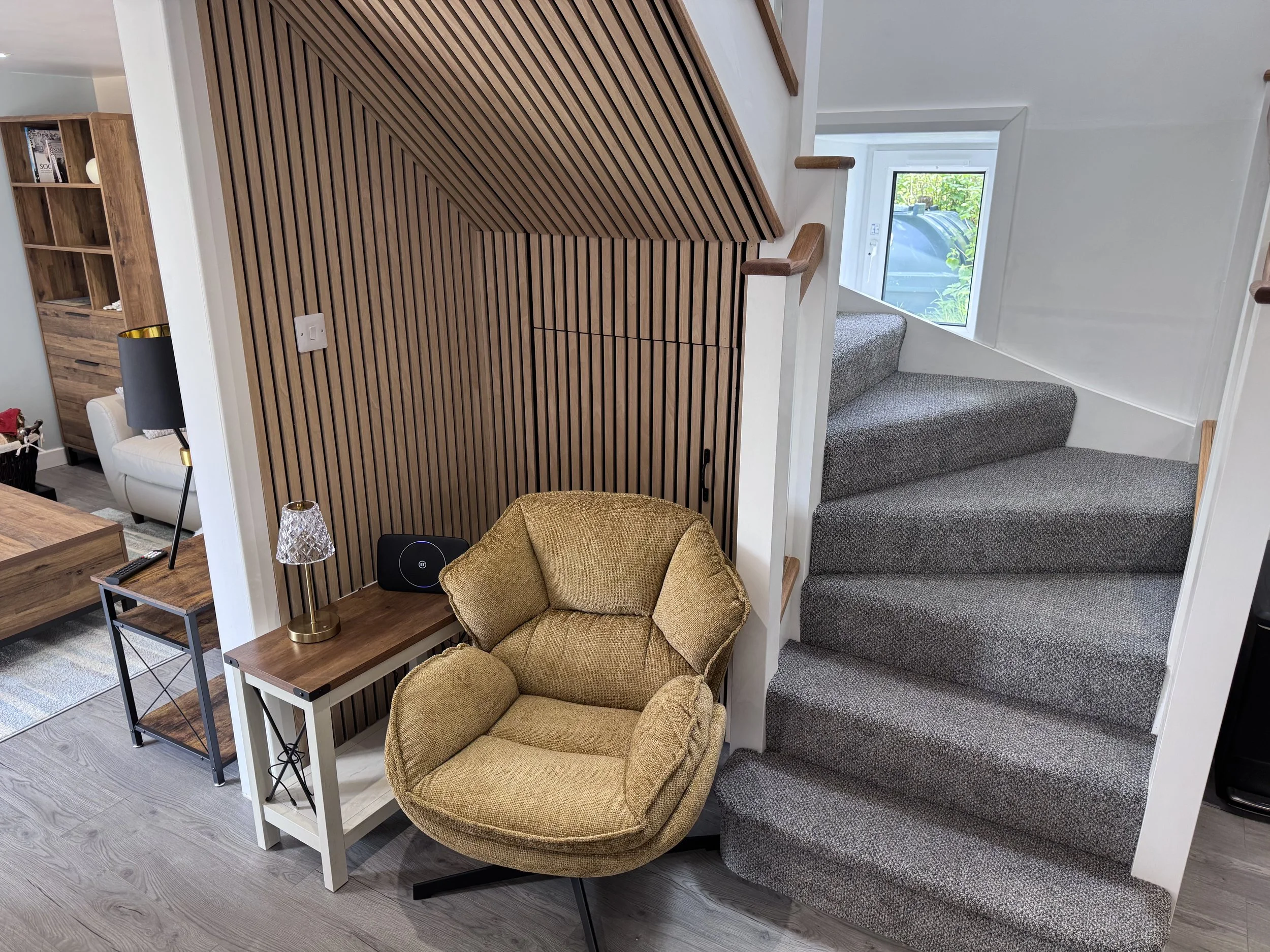 Interior of a modern home with a wooden dividing wall, a staircase with gray carpeting, a window showing greenery outside, a white armchair, a wooden shelving unit, a small side table, a table lamp, and a decorative glass object.