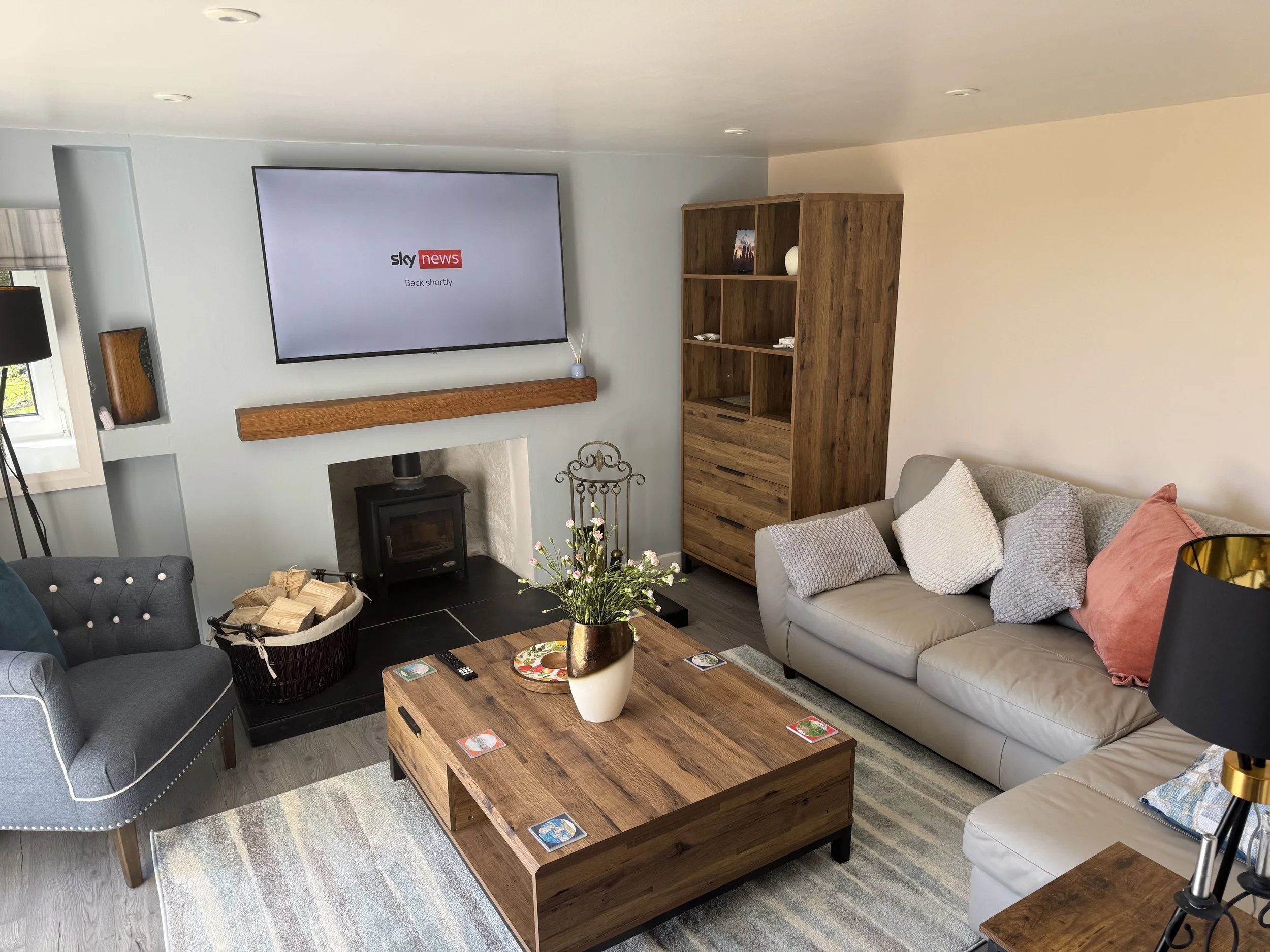 Living room with a wall-mounted TV displaying 'sky news back shortly', a wood stove, a wooden coffee table, a gray couch with pillows, a magazine on the table, a basket of firewood, a flower vase, a wooden shelving unit, a window with a curtain, and a tripod with a camera.