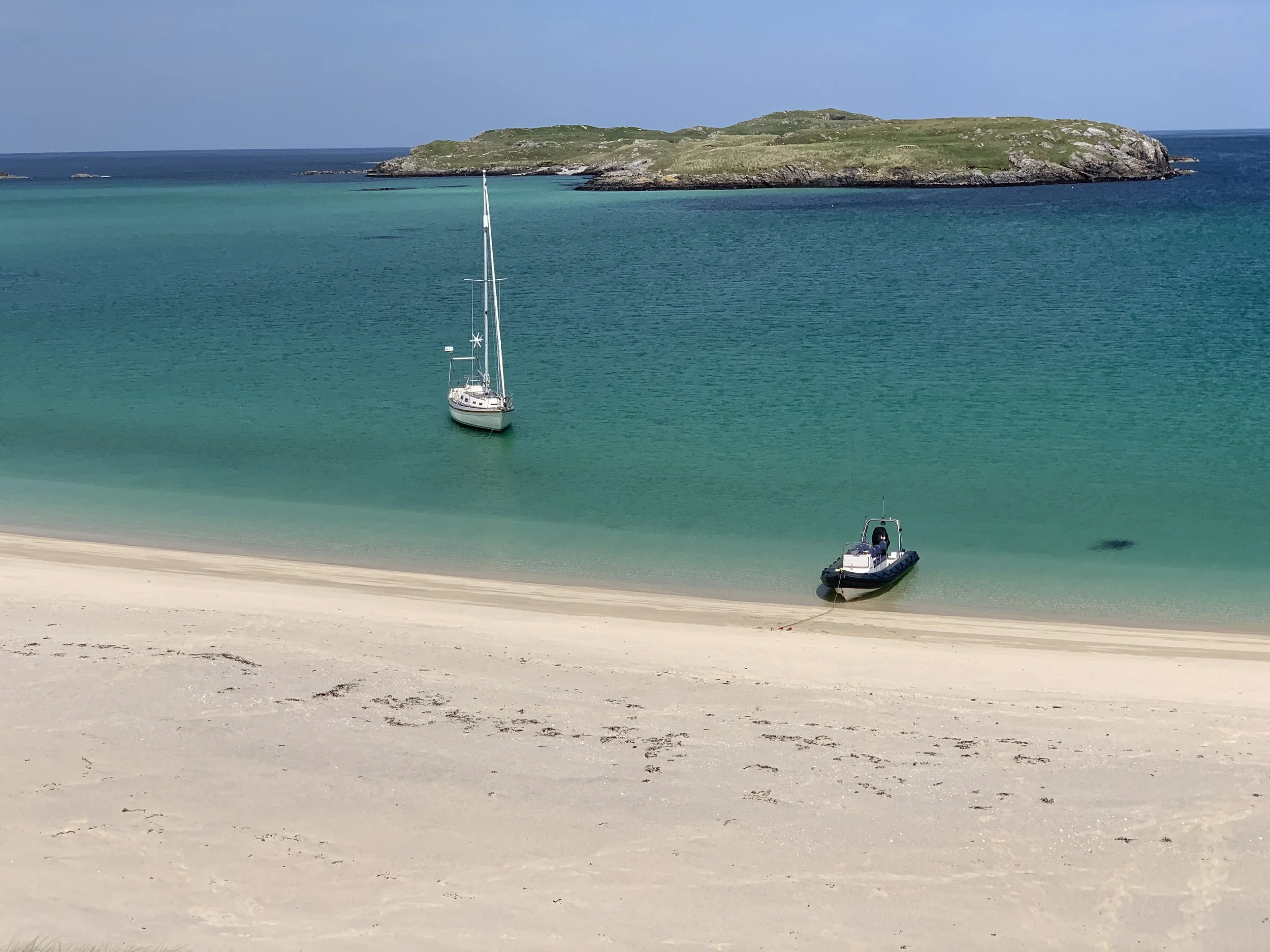 A sandy beach with a sailboat anchored in turquoise water near a rocky island with green grass. A motorboat is on the beach in the foreground.