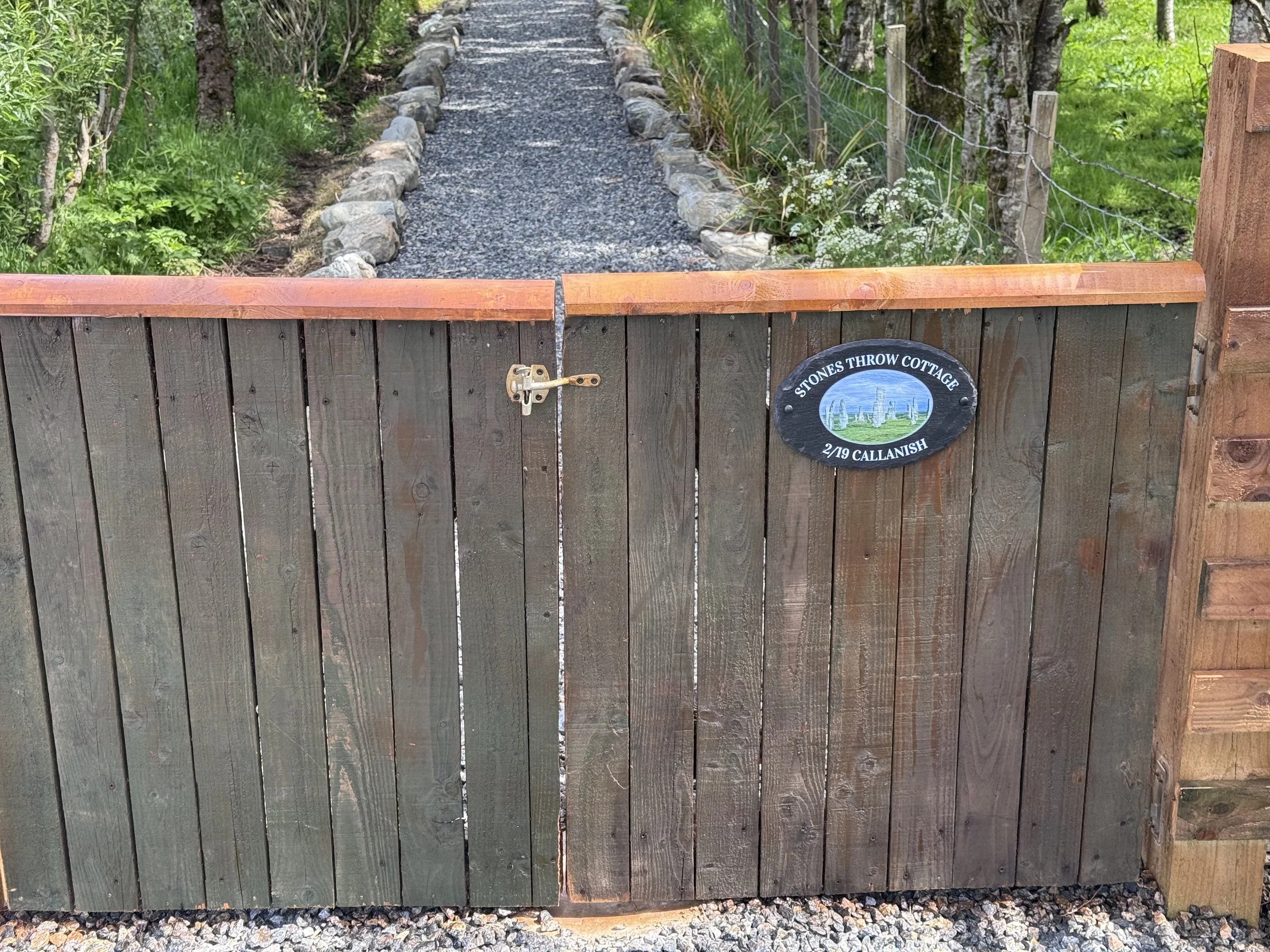 A wooden gate with a lock, part of a fence, with a gravel pathway leading into a lush green garden or park area.