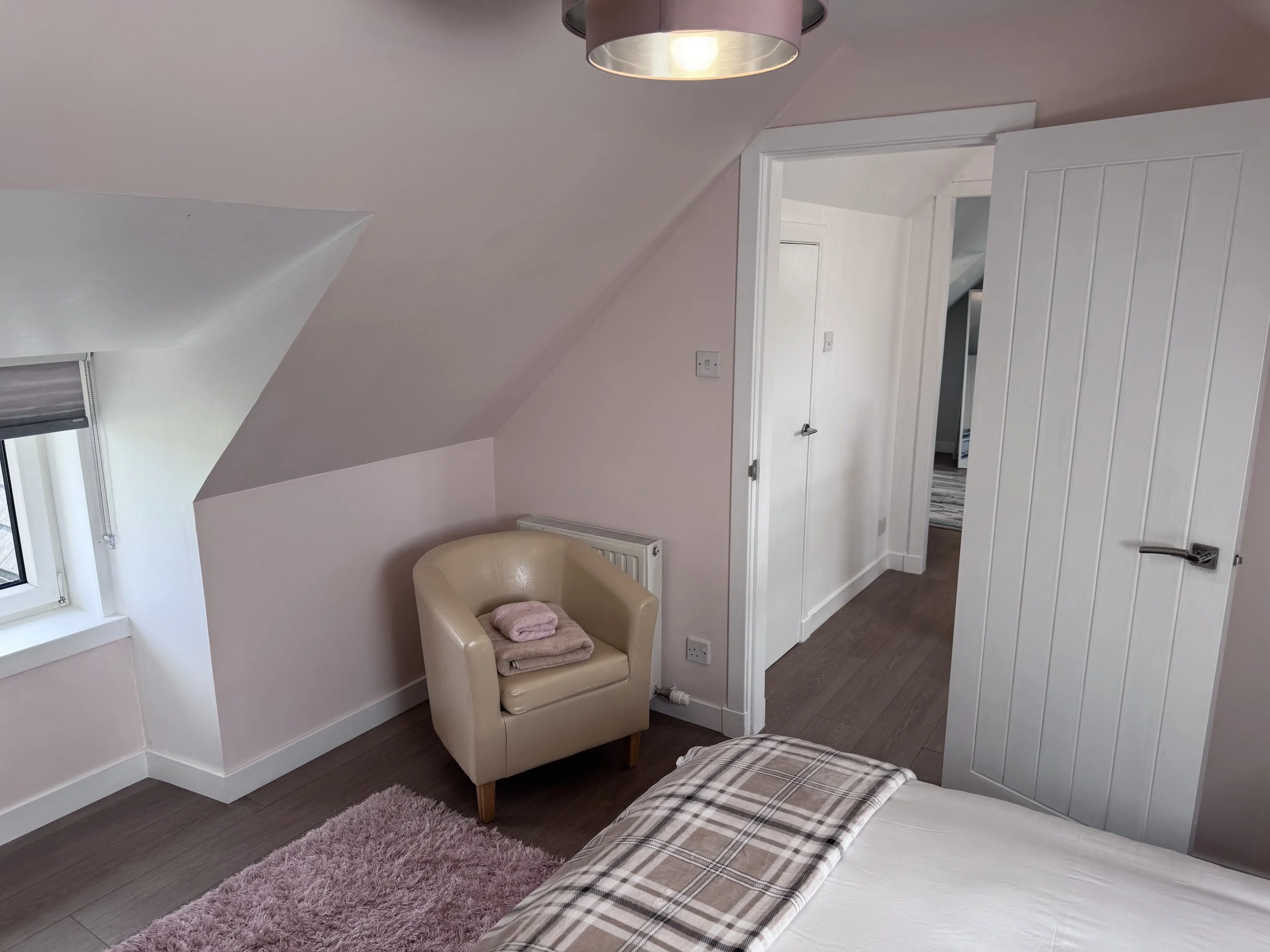 A cozy bedroom with a slanted ceiling, a window with closed blinds, a beige armchair with folded towels on it, a fluffy pink rug, a bed with plaid bedding, and an open door leading to another room.